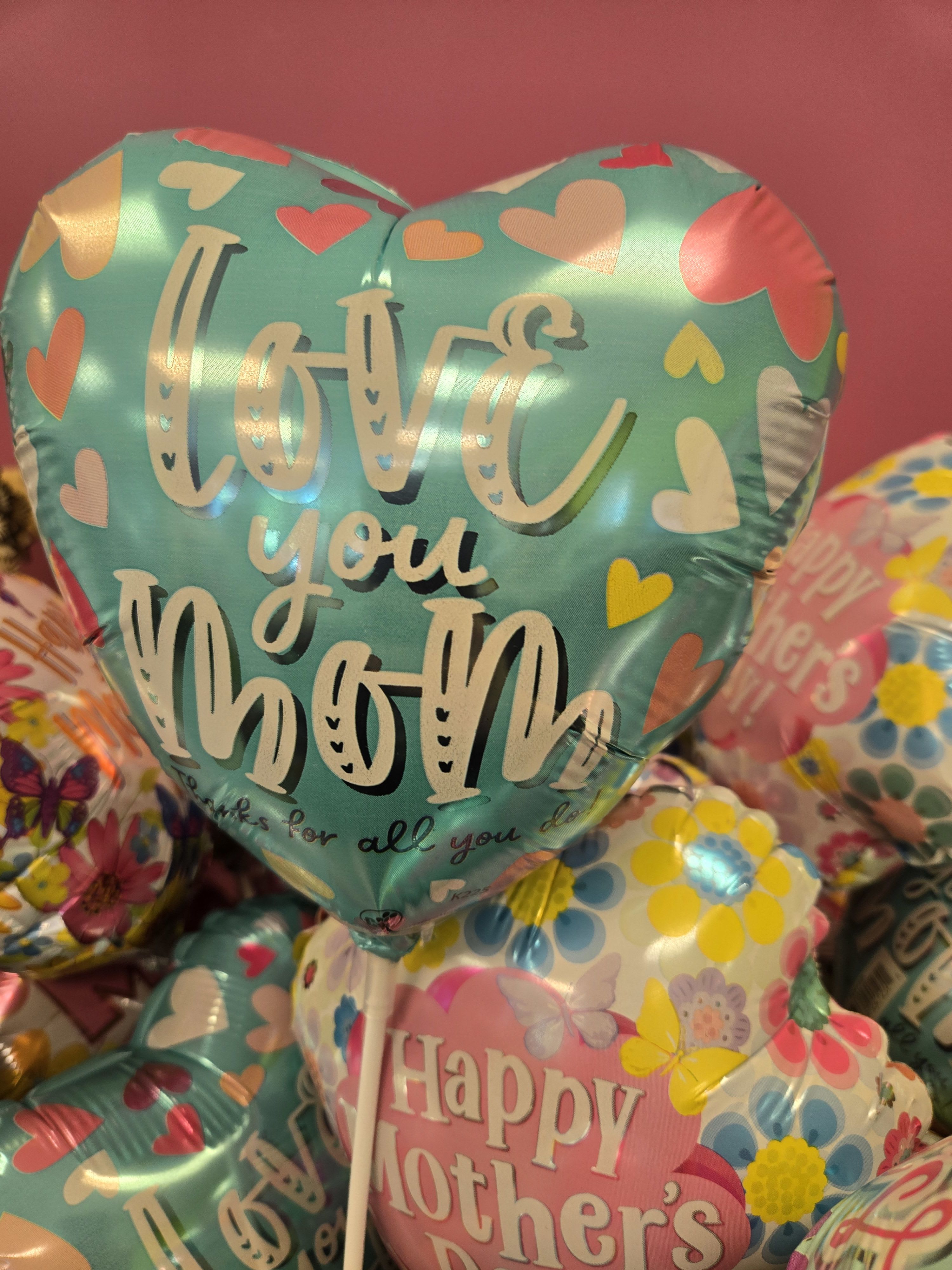 assorted mini mylars - A glossy cluster of heart-shaped foil balloons that says "Love you Mom" in bold teal lettering creates an immediate Mother's Day focal point - a cheerful Mother's Day balloon bouquet that feels affectionate and celebratory. This assortment of mini mylars is perfect for honoring mom, grandma, or any special mother-figure who deserves a bright, joyful surprise that reads clearly from across the room.  The set includes multiple heart and floral-patterned foil balloons in pink, blue, yellow, and white, arranged on balloon sticks for a tidy, gift-ready display that fits atop a table, gift basket, or dessert table. The shiny surfaces catch the light and overlap for a full, festive look without needing helium; each mini mylar is secured on a stick so the cluster keeps its shape and makes a polished presentation.  Ideal for Mother's Day, a birthday, or a thoughtful "just because" moment, this bouquet sends a warm, loving message that will make her feel cherished. Easy to place with included sticks and ready to pair with flowers or a cake, it's a simple way to create a memorable centerpiece or add an extra pop of celebration to your gift.