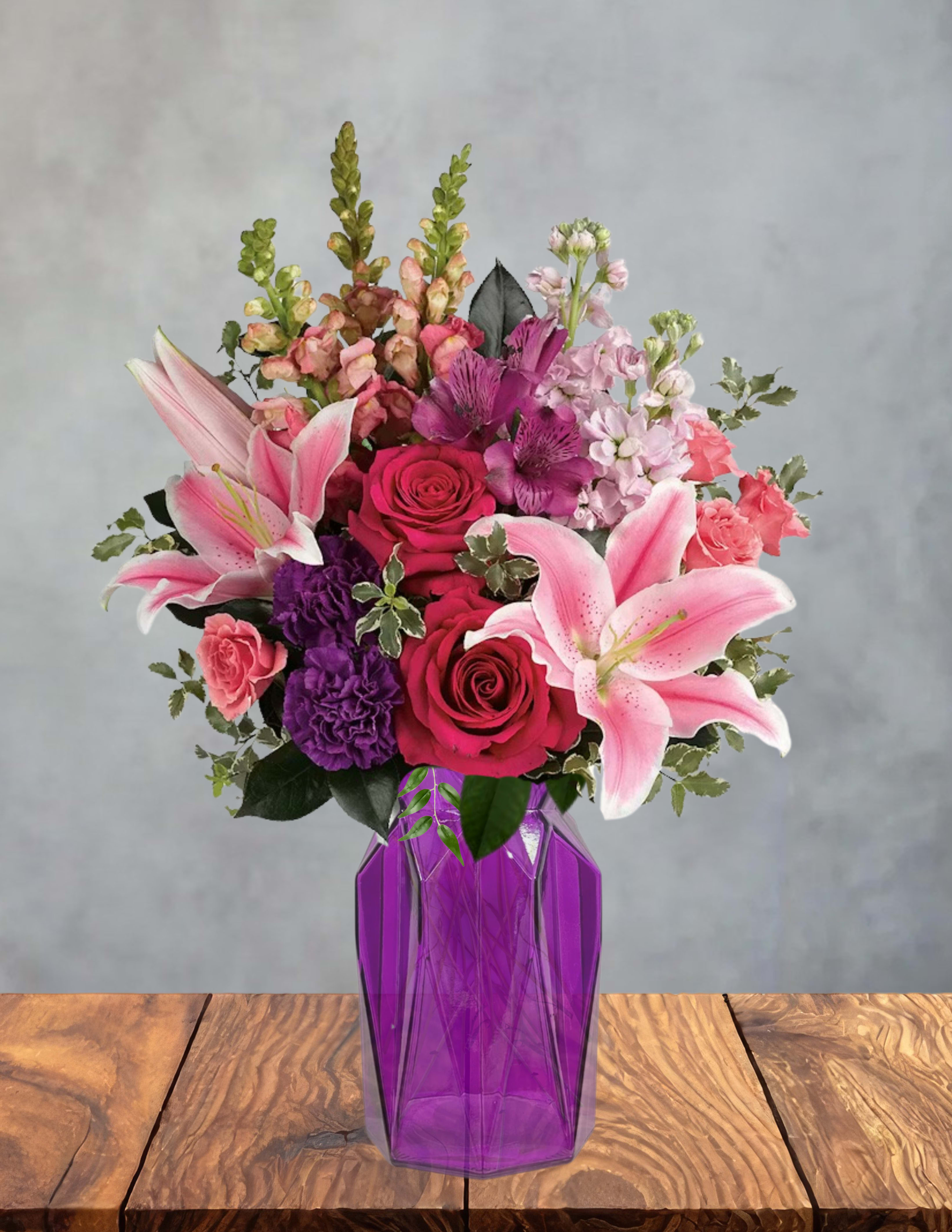Purple stardust  - Bright, lush pink lilies reach outward over a garden of hot pink and red roses, purple carnations, and pale pink snapdragons-an arrangement made for birthdays, anniversaries, or any romantic moment when you want the person you're thinking of to feel celebrated. As a flower arrangement for love and romance or a cheerful thank-you gift, Purple stardust combines saturated pinks and violets that read both luxurious and joyful, sending a clear message of warmth and affection.  Arranged upright in a faceted purple glass vase, the design layers large, fragrant lilies with clustered roses and ruffled carnations while snapdragons add vertical texture and softness. The composition is full and abundant, roughly tabletop height and sized to make an immediate visual impact without overwhelming a space. The purple vase ties the palette together and makes this bouquet an easy centerpiece that brightens a room.  Perfect for surprising a partner, honoring a friend's birthday, or saying thanks with flair, the recipient will feel genuinely seen by the bold color mix and generous blooms. Place in fresh water and trim stems slightly every few days to extend vase life; order now for local delivery to ensure peak freshness on the special day.