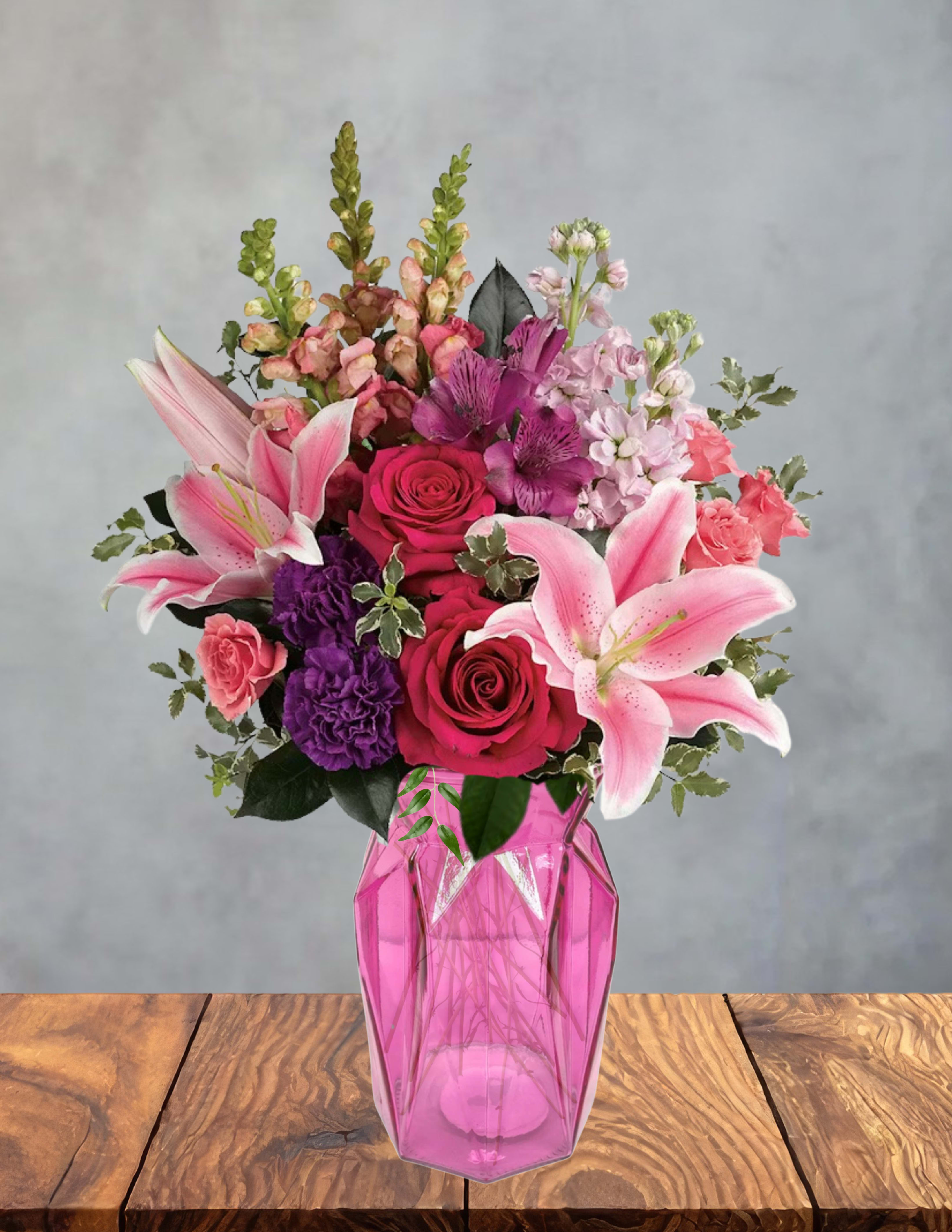 Pink stardust - Big, blousy pink lilies take center stage in this Pink stardust flower arrangement, surrounded by hot pink and red roses, purple carnations, and delicate pale pink and lavender filler blooms-perfect as a birthday bouquet, anniversary flowers, or a romantic surprise. The tall snapdragon spikes add elegant height while the lush mix of saturated pinks, purples, whites, and fresh greenery creates a joyful, celebratory impression that will make the person you're thinking of feel truly seen.  Arranged in a faceted pink glass vase, the jewel-toned container amplifies the arrangement's glossy, modern look and keeps stems neatly gathered. The composition reads full and abundant with varied textures-from the lilies' smooth petals to the ruffled carnations and the delicate alstroemeria-so it works as a centerpiece or a standout gift. Ideal for everyday cheer or special moments, this arrangement arrives ready to display; change the water and trim stems every few days to extend freshness and enjoy the colors longer.