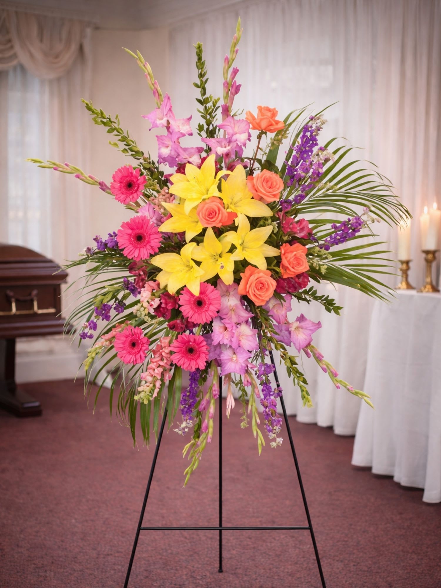 Uplifting Sendoff Standing Spray  - Uplifting Sendoff Standing Spray is a vibrant and joyful floral tribute designed to celebrate a life beautifully lived. This striking standing spray features radiant yellow lilies, bright pink gerbera daisies, coral roses, and soft pink gladiolus accented with purple stock and lush tropical greenery. The colorful palette of yellow, pink, coral, and lavender creates a warm and uplifting display that honors cherished memories while bringing comfort to family and friends.  Expertly designed by the floral artists at Floral Fabulous, a trusted local florist in Round Rock TX near Austin, this standing spray is a perfect tribute for celebration-of-life ceremonies, funeral services, and memorial gatherings. Yellow lilies symbolize hope and remembrance, while gerbera daisies represent cheerfulness and gratitude for a life well lived. Roses and gladiolus add elegance and heartfelt beauty to this expressive arrangement.  Displayed on a standing easel, the Uplifting Sendoff Standing Spray creates a beautiful and meaningful presence during memorial services. Each arrangement is handcrafted with fresh premium flowers to ensure a stunning tribute that reflects love, remembrance, and celebration.  Floral Fabulous proudly provides professional funeral flower delivery in Round Rock, Pflugerville, Georgetown, and throughout the greater Austin Texas area. Uplifting Sendoff Standing Spray is a thoughtful way to celebrate a life, express sympathy, and honor lasting memories with warmth and color.