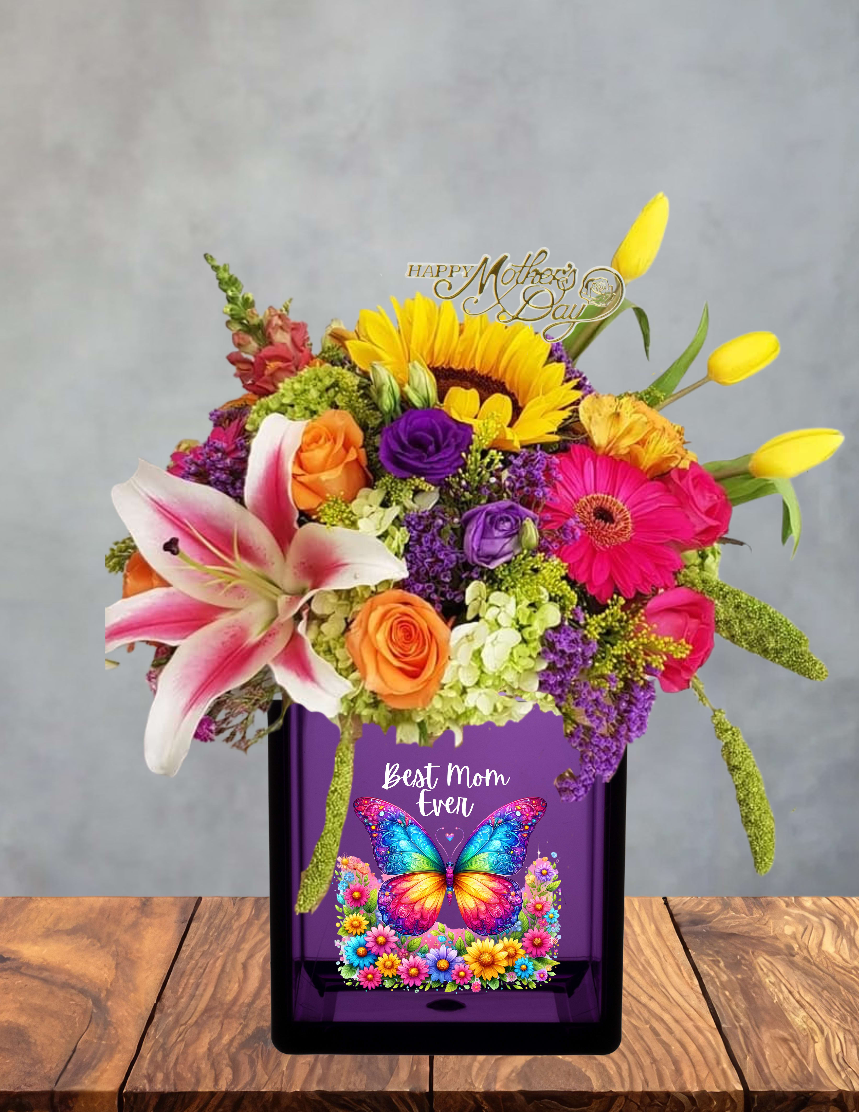 Flutter and bloom  - A vivid, garden-fresh arrangement designed to make Mom feel cherished the moment she sees it-this mixed-color flower arrangement is perfect as a Mother's Day bouquet or a celebratory gift for a mom's birthday or thank-you moment. Bright sunflowers and pink gerbera daisies create an immediate burst of sunshine, while orange and purple roses plus a pink-and-white lily add depth and a soft, fragrant focal point that reads joyful and heartfelt to the recipient.  The design is full and abundant: a large sunflower anchors the mix alongside blooming roses, a prominent lily, hydrangea textures, snapdragons, yellow tulip buds, and purple filler florals for height and movement. It arrives in a glossy purple vase with a decorative butterfly plaque that reads "Best Mom Ever," giving the gift a personalized, keepsake quality. Colors range from sunny yellow and warm orange to soft pinks and vivid purples for a lively, celebratory palette.  Made to feel like a thoughtful, confidence-inspiring gift, this arrangement is the kind of bouquet that will make her feel truly seen and celebrated. Order with standard care: keep blooms in fresh water, trim stems slightly every few days, and place away from direct heat to extend vase life-perfect for brightening a kitchen table, bedside, or living-room centerpiece.