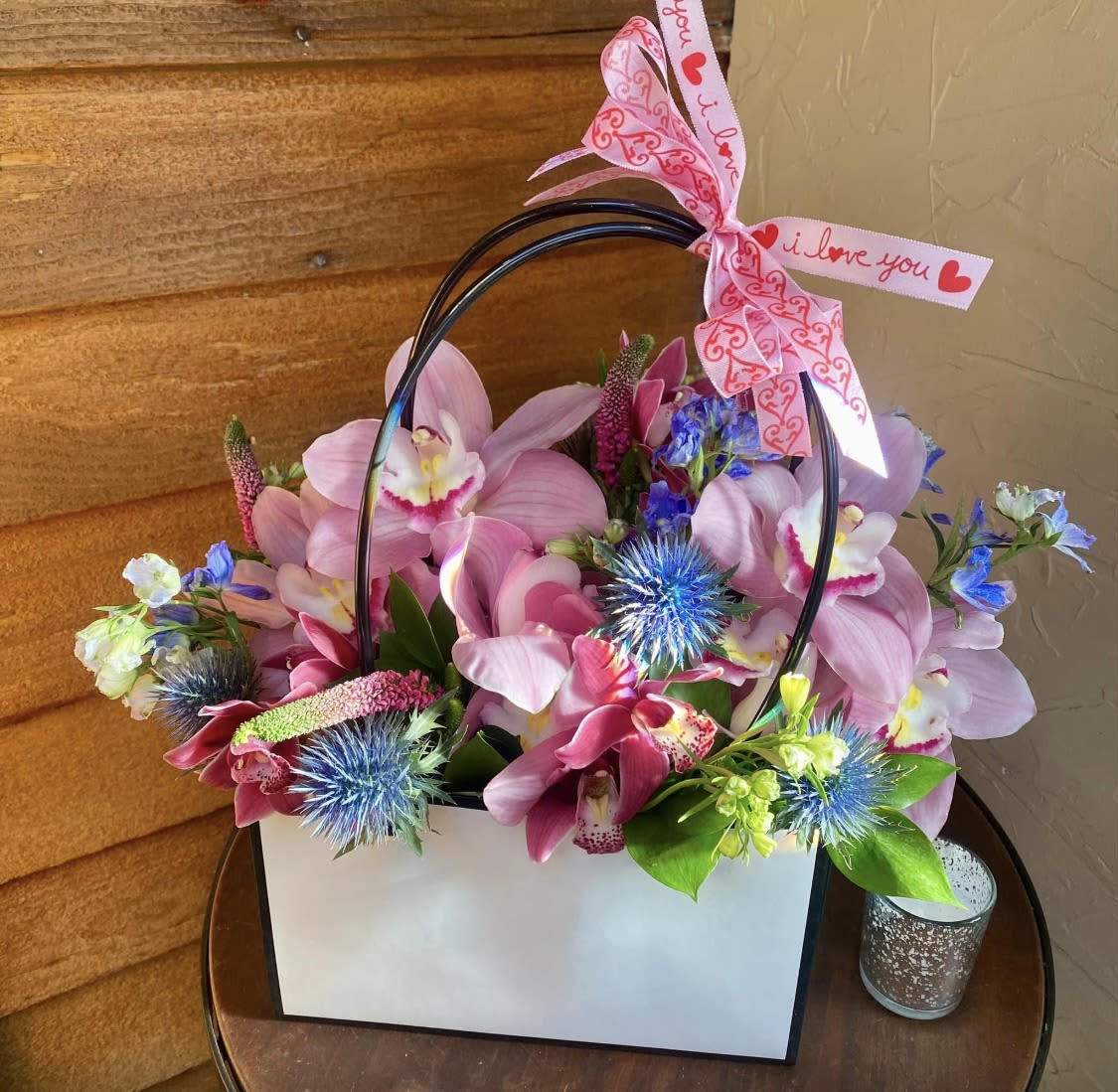 Pink orchid arrangement in a white gift bag with a heart ribbon