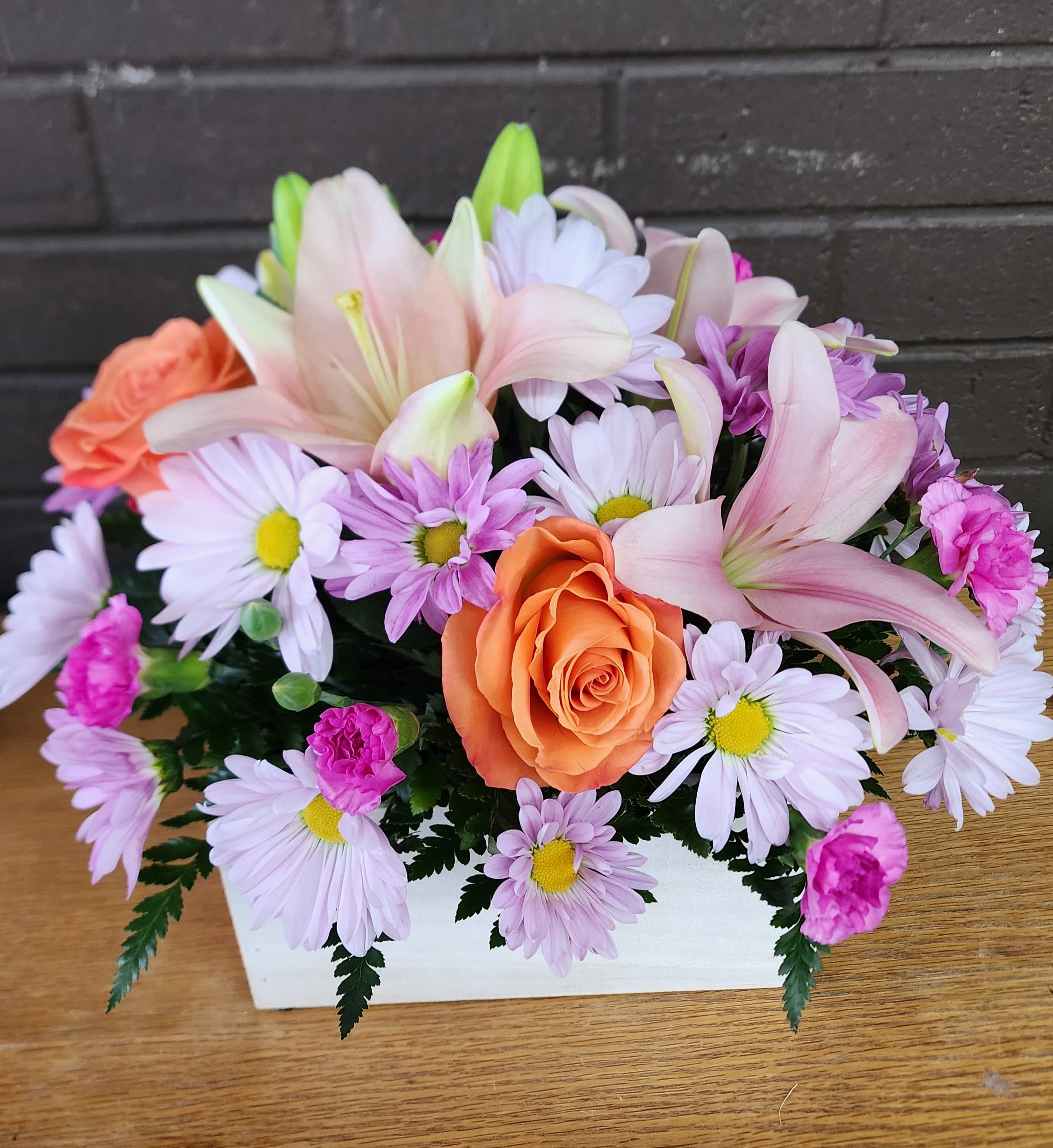 Love and Light - Pale pink lilies and warm orange roses create a cheerful, springlike first impression in this flower arrangement for birthdays, thank-you gestures, or a thoughtful thinking-of-you surprise. The low white rectangular box keeps the composition intimate and easy to display, so the person you're gifting will feel genuinely seen by a bright, romantic burst of color.  Closely packed lavender daisies and bright pink carnations fill the design for a full, rounded silhouette with soft pastels and a lively orange accent. The mix of lilies, roses, daisies, and carnations offers varied textures - silky petals, ruffled carnation edges, and button-like daisy centers - that read fresh and abundant on a tabletop or desk. The compact size in a white container makes it a simple care arrangement: trim stems, refresh water, and keep out of direct heat to prolong blooms.