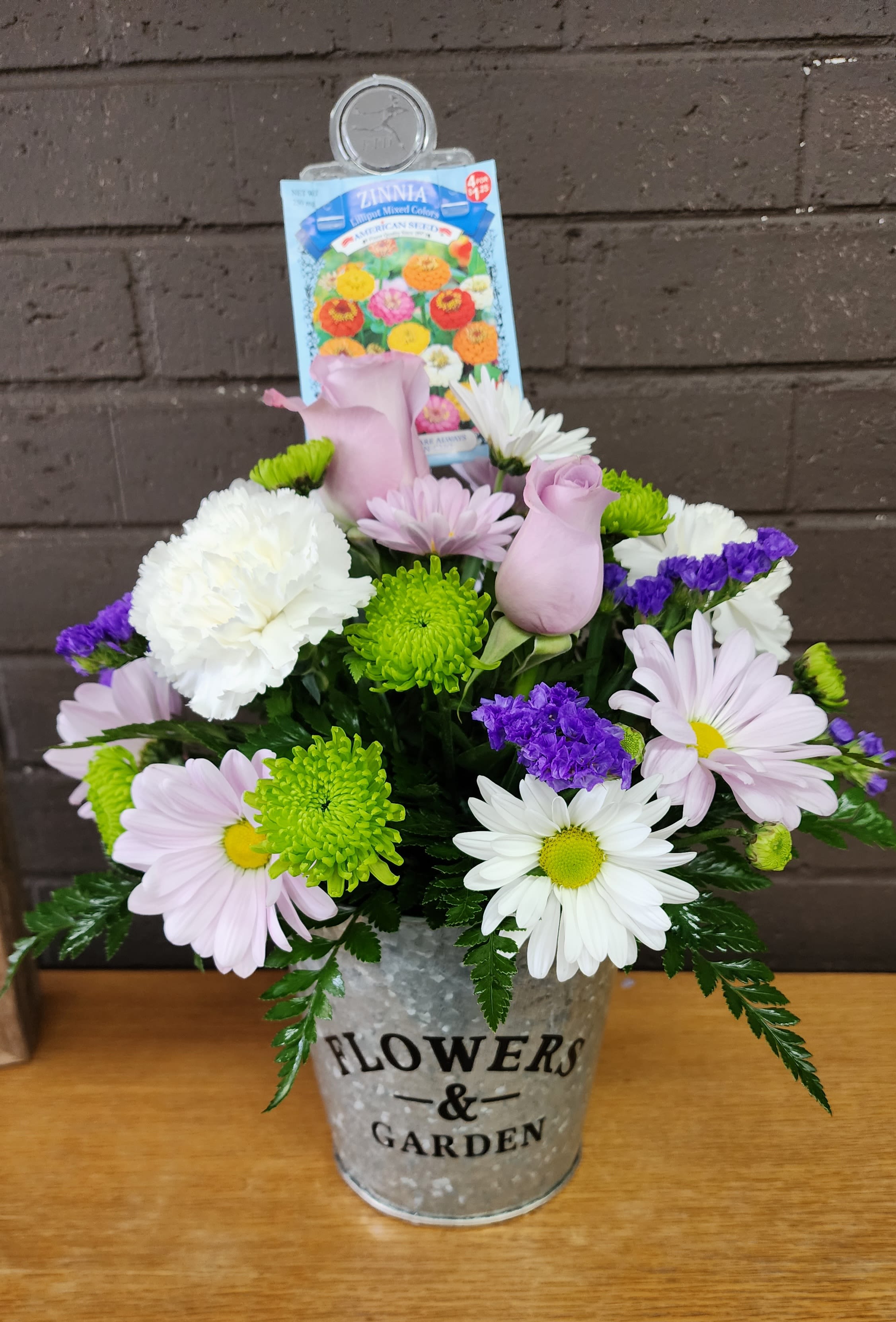 Flowers and Garden - Pale pink roses and lavender-and-white daisy-like blooms mingle with bright green and purple chrysanthemums in a galvanized metal bucket-an upbeat, garden-style flower arrangement perfect for a birthday bouquet, housewarming gift, or just-because surprise. The lively color mix and rounded, closely layered composition create an immediate cheerful impression that will make the person you're thinking of feel noticed and celebrated.  This full arrangement includes roses, daisies, and chrysanthemums arranged densely for a lush, textured look; the galvanized bucket gives a casual, modern farmhouse vibe and makes the piece ready to display. A flower seed packet tucked into the back reinforces the spring-planting theme and adds a thoughtful keepsake. Ideal for tabletops, entryways, or giving to someone starting a new home, this arrangement arrives ready to brighten a room-just add fresh water and enjoy.