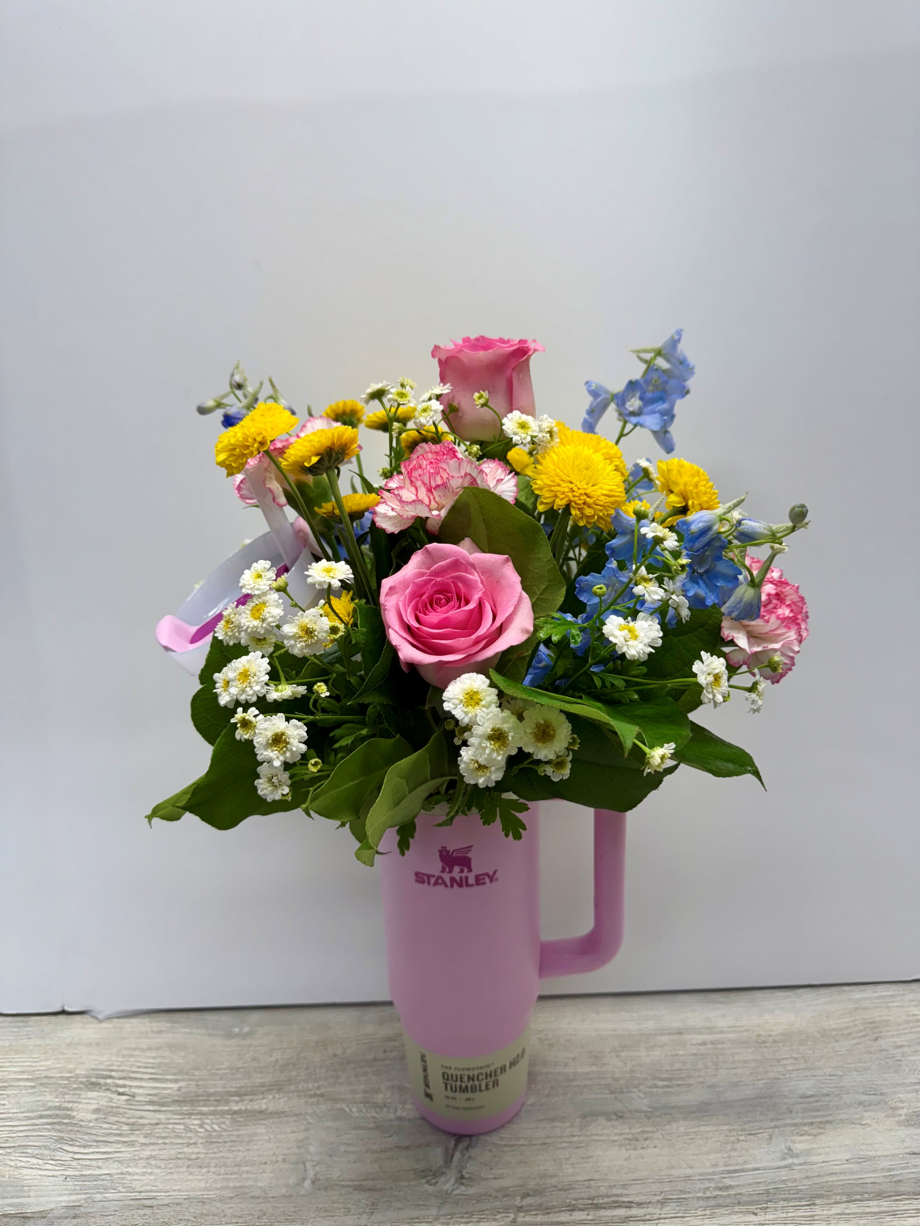Mixed bouquet of pink roses, carnations, yellow mums, and blue blooms arranged in a pink tumbler.