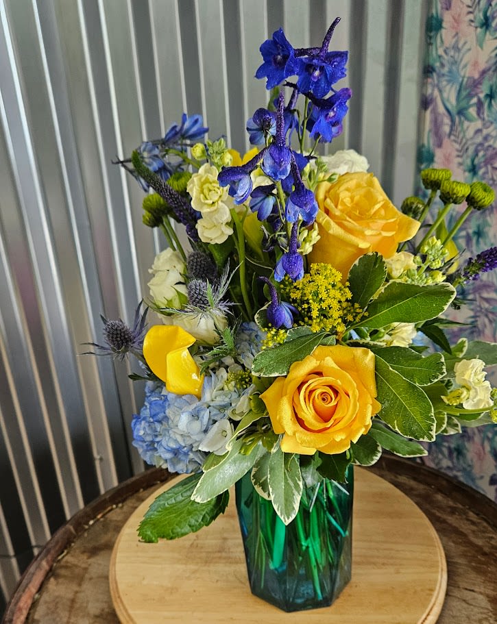 Ocean Sunrise - Experience the refreshing beauty of an Ocean Sunrise with this radiant floral arrangement. Inspired by the tranquil meeting of sea and sky at dawn, this bouquet features sunny yellow roses, vivid blue delphiniums, soft blue hydrangeas, creamy spray stock, and playful accents of thistle and greenery. Nestled in a striking Blue green glass vase, this arrangement brings a splash of coastal charm and uplifting energy to any space. Perfect for Get Well, birthdays, celebrations, or simply adding a touch of sunshine to your day. Highlights: Features yellow roses, blue delphiniums, hydrangeas, spray stock, thistle, and lush greenery Arranged in a faceted blue-green glass vase Handcrafted with care and attention to detail