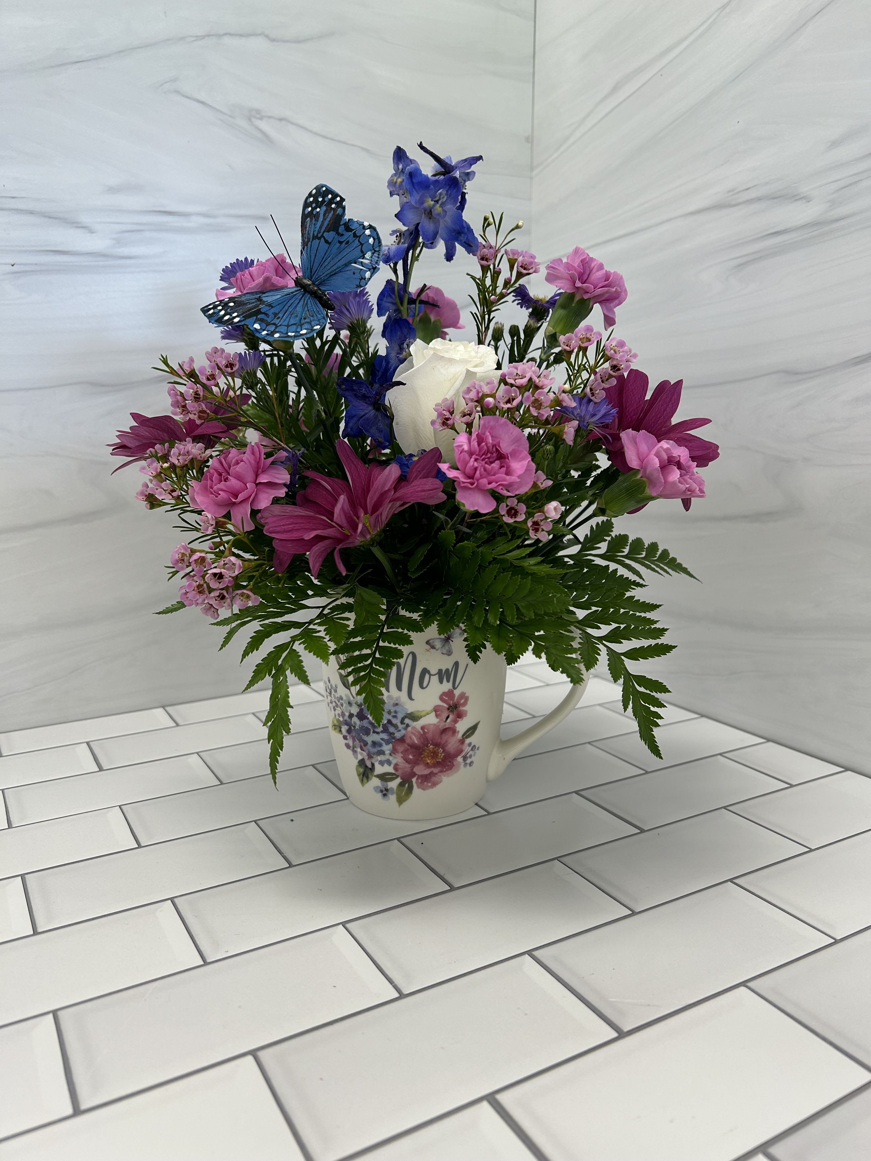 A Hug for Mom - The perfect little arrangement to let mom know you care.  This mug arrangement features daisies, mini carnations, delphinium, spring asters and a white rose. Bouquet will be delivered approximately as pictured.