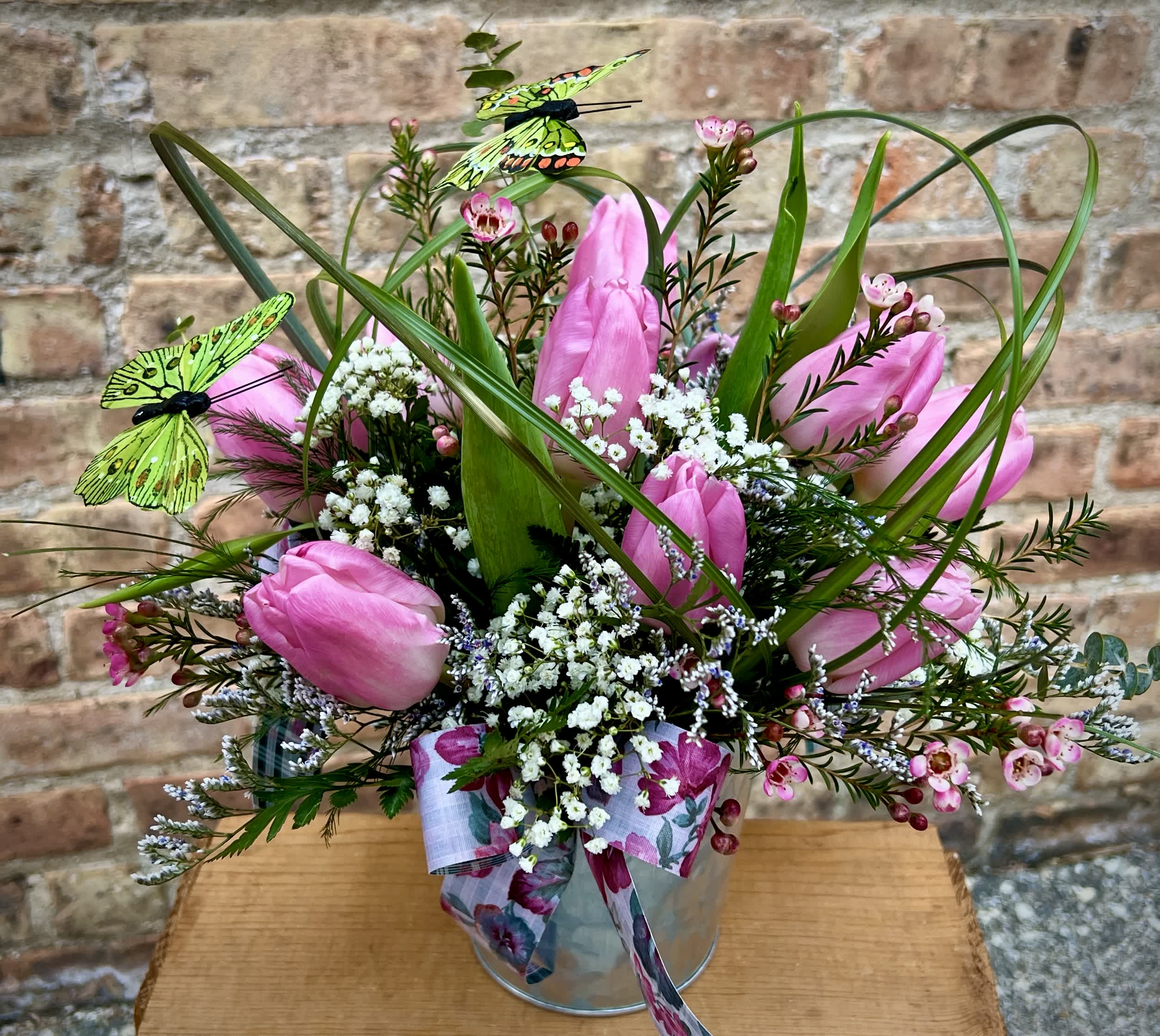 Pink tulip arrangement with white filler flowers, grass loops, and decorative butterflies in a metal pail.