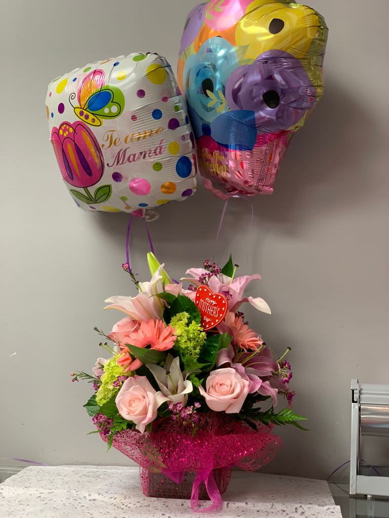 CHERISHED BLOOMS  - Soft pink roses, fragrant lilies, and cheerful gerbera daisies come together in a compact Mother's Day bouquet designed to make her feel truly cherished. This birthday or Mother's Day gift arrangement pairs the mixed-color floral spray with two helium foil balloons-one with a Mother's Day message and one playful polka-dot print-so the person you're celebrating sees the love before they even lift the lid of the decorative basket.  The flowers sit in a festive pink mesh wrap inside a woven basket, layered with lavender and white blooms, bright green accent buds, and delicate filler flowers for texture and depth. A heart pick adds a tender finishing touch and the two balloons float above for instant visual impact. This arrangement is mid-sized and ready to display on a table or counter; light floral care-fresh water and a cool spot away from direct sun-will help the roses and lilies last longer.  Perfect for moms, grandmothers, or any woman you want to honor, this arrangement reads as affectionate and celebratory-an easy way to say "thank you" or "I love you" without words. Order with same-day delivery where available to ensure the balloons and blooms arrive bright and ready to make her day.