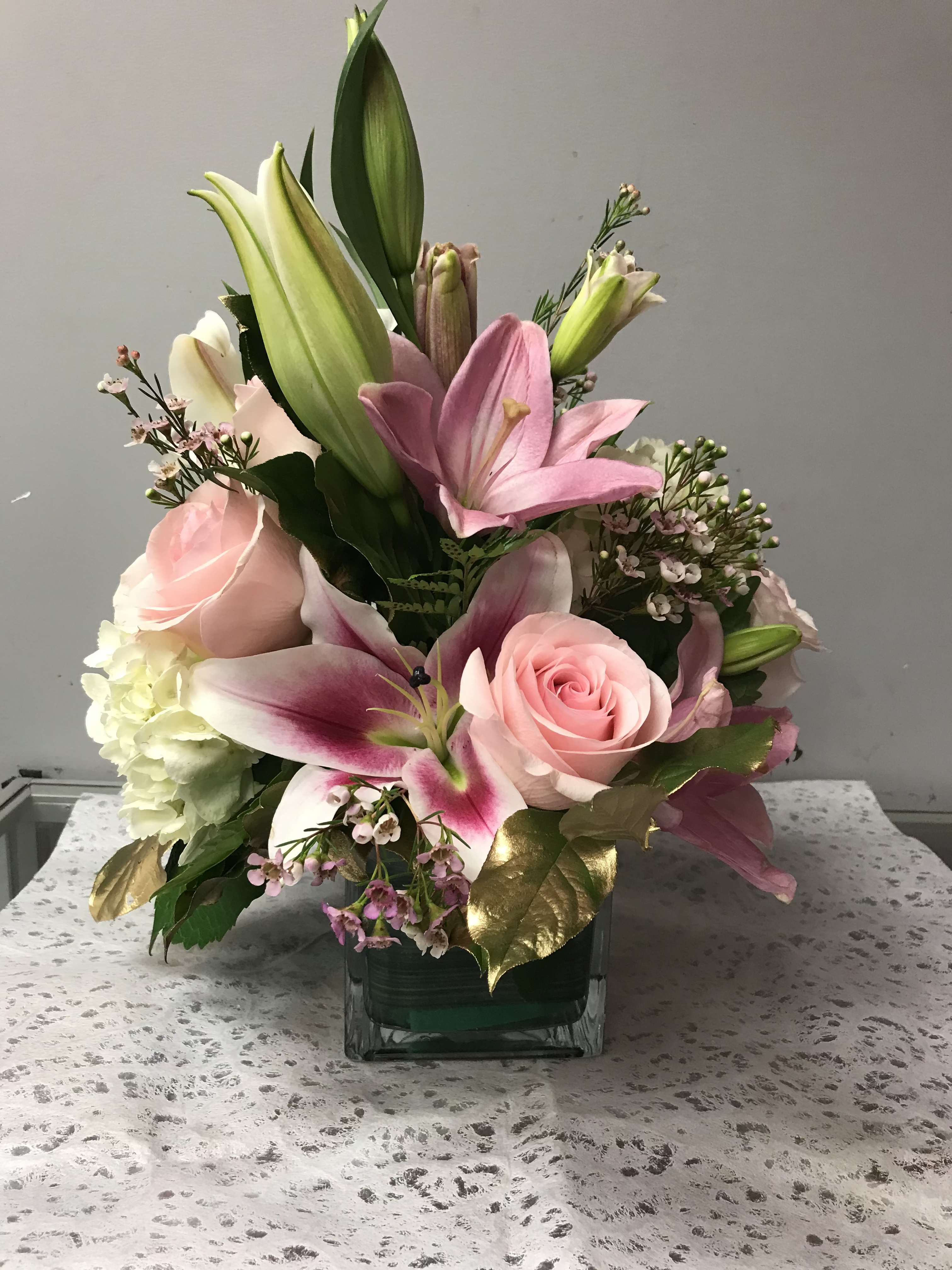 GIFT DELICATE  - Soft pink lilies and pale pink roses sit above a dense cloud of white hydrangea in this compact flower arrangement-perfect as a birthday bouquet, anniversary gift, or a just-because surprise for the person you're thinking of. The lilies' tall buds rise gracefully over the tightly layered cluster, creating an upright, sculpted silhouette that feels both romantic and refined for Mother's Day or any special moment.  Creamy whites and soft pinks are warmed by a few metallic gold leaves for a subtle, formal touch. Arranged in a square glass vase, the design is compact and table-ready, measuring as a medium-sized centerpiece ideal for a bedside table, dining room, or office. The mix of lilies, roses, hydrangea, and delicate filler blooms gives a lush, textured look-she'll feel truly celebrated when this arrives.