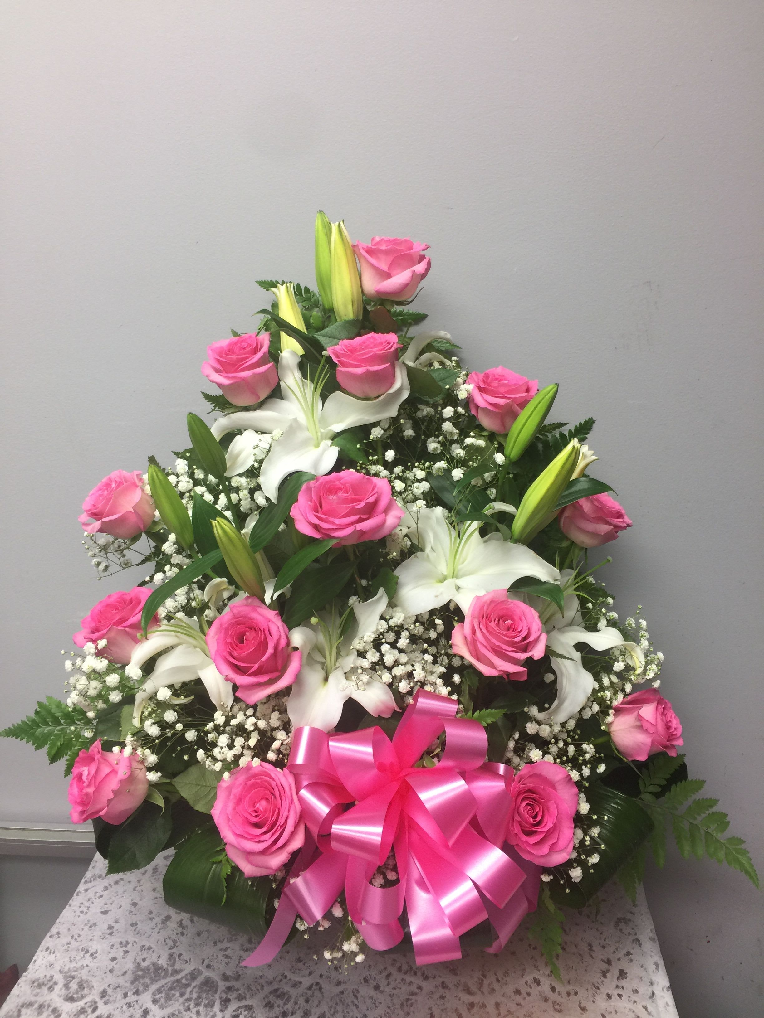 LOVELY MOM ROSES  - Bright pink roses paired with creamy white lilies create a lush, feminine birthday bouquet or anniversary arrangement that reads as both romantic and celebratory. The tiered, rounded design gives this floral gift a formal presence-perfect for making the person you're thinking of feel honored and truly seen on a special day or as a heartfelt thank-you.  Clusters of delicate baby's breath and small white filler flowers tuck between the roses and lilies, adding soft, airy texture against deep green foliage. A large shiny pink satin ribbon bow anchors the base, giving the piece a polished, gift-like finish. The color story-pink, white, and green-plus the mix of velvety rose petals and sculpted lily blooms creates a fragrant, tactile bouquet that reads as joyful and elegant.