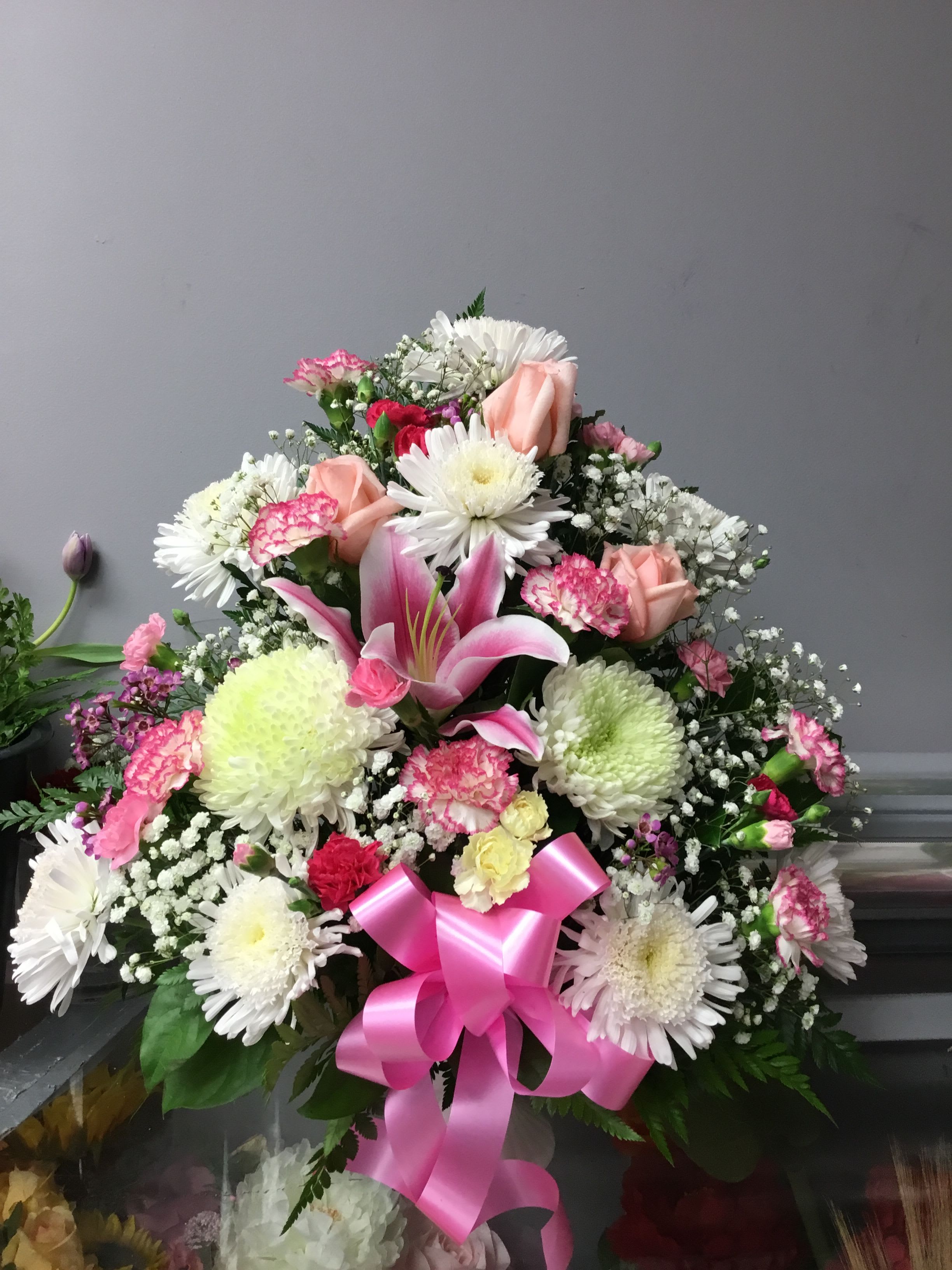 MOMS DELIGHT  - A lush, rounded bouquet of pink and white blooms that reads as a warm, celebratory Mother's Day or birthday arrangement-this flower arrangement is designed to make the person you're thinking of feel cherished. The mix of prominent pink lilies and pale roses gives a soft, romantic focal point while the carnations and chrysanthemums add body and longevity; small sprays of baby's breath lend an airy, delicate texture. The overall palette of pink, white, and mixed pastels makes it equally fitting for a thank-you, anniversary, or sympathy gesture.  Generous in size with a dense, layered shape, the arrangement is finished with a bright pink satin ribbon bow at the base for a polished, feminine touch. Expect a blend of long-lasting varieties (roses, carnations, chrysanthemums) alongside showy lilies and daisies that create visual contrast and gentle scent. Made to present beautifully on a table or mantle, this bouquet is the kind of gift that helps someone feel truly seen-choose it when you want to celebrate, comfort, or honor a loved one with timeless, bloom-filled elegance.