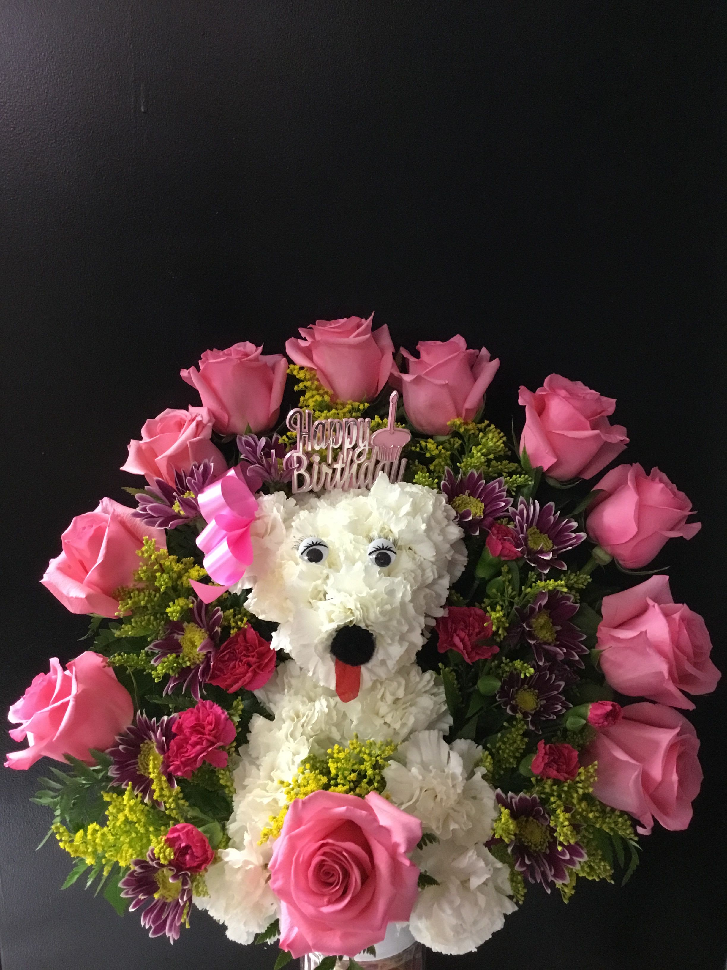 PRECIOUS PUP  - A whimsical dog-shaped flower arrangement made for birthdays, this playful bouquet turns flowers into a keepsake moment the person you're celebrating will smile at. The birthday topper and novelty facial details-the little plastic eyes, black nose, and red tongue-make the arrangement feel like a surprise companion, while the pink, white, purple and yellow palette reads cheerful and festive for a birthday gift.  White blooms form the pup's face and body, bordered by pink roses with magenta carnations, purple daisy chrysanthemums and sunny yellow filler flowers layered in for texture and contrast. Decorative ribbon finishes the design and reinforces the celebratory look; proportions are compact and tabletop-friendly so the floral pup fits easily on a counter, desk, or side table.  Perfect for a child, a dog lover, or anyone who appreciates a lighthearted birthday gesture, this arrangement sends a playful, affectionate message-she'll feel seen and delighted. Order with standard delivery for a ready-to-display surprise; the included novelty pieces are attached for a long-lasting presentation, and basic care-fresh water and a cool spot away from direct sun-will help the blooms last.