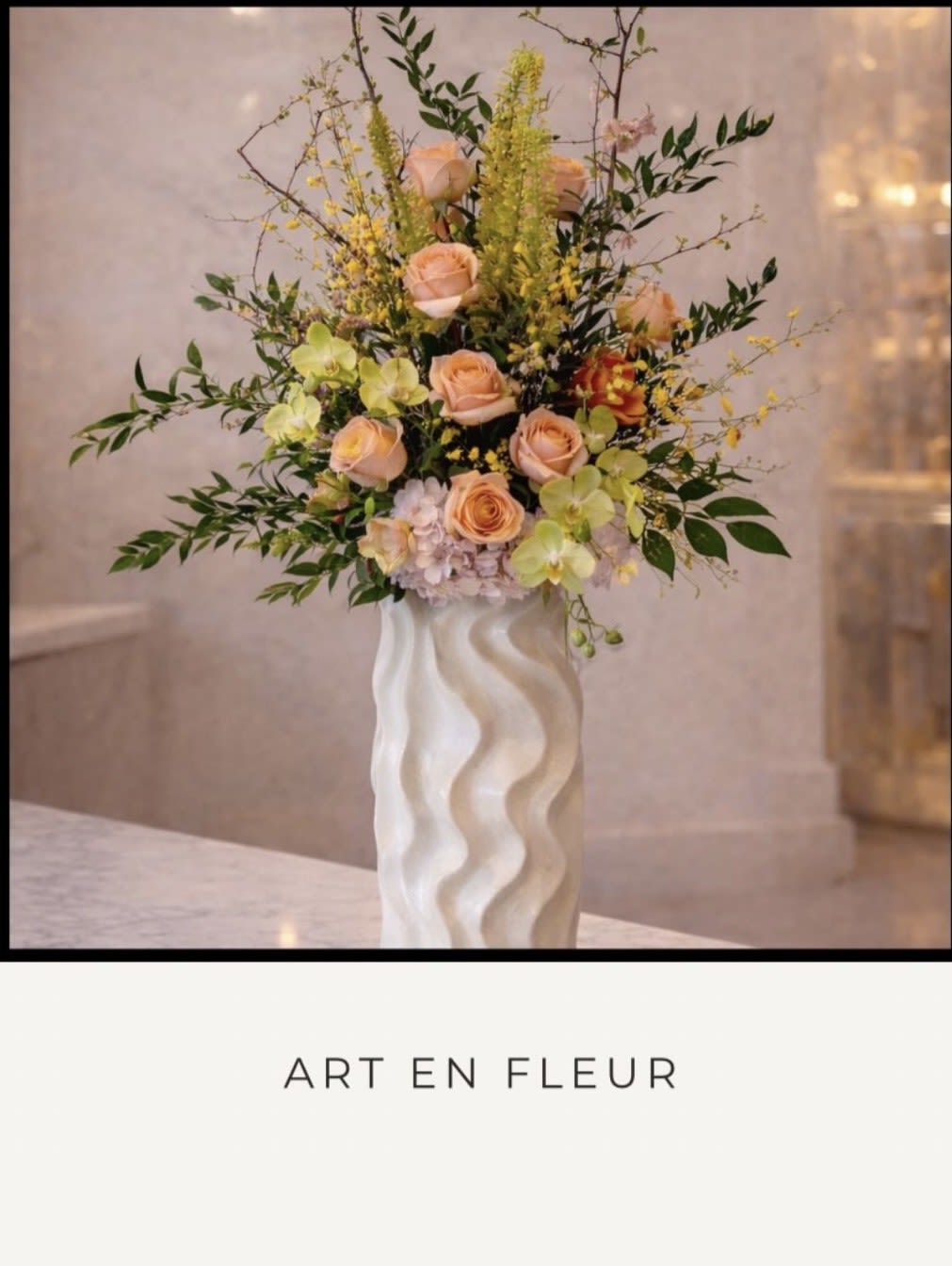 Art En Fleur - Peach roses and pale green orchids rise from a soft lavender hydrangea base in a tall, sculptural display-an elegant flower arrangement perfect for birthdays, anniversaries, housewarmings, or a heartfelt thank-you. The vertical, garden-inspired silhouette feels contemporary yet romantic, the kind of bouquet that makes the person you're thinking of feel genuinely seen and celebrated.  This arrangement combines roses, orchids, hydrangeas and delicate yellow filler stems arranged with dark, branching foliage for height and movement. The flowers sit in a white ceramic vase with a wavy, sculpted surface that adds modern contrast and works as a decorative piece long after the blooms soften. Colors include warm peach and blush, fresh green, soft lavender and creamy yellow accents for a balanced, refined palette.  Ideal for dressing an entry table, brightening a living room, or marking a special milestone, this composition arrives ready to display. For best longevity, place in a cool spot away from direct sunlight and refresh the water every couple of days to keep the blooms looking their freshest.