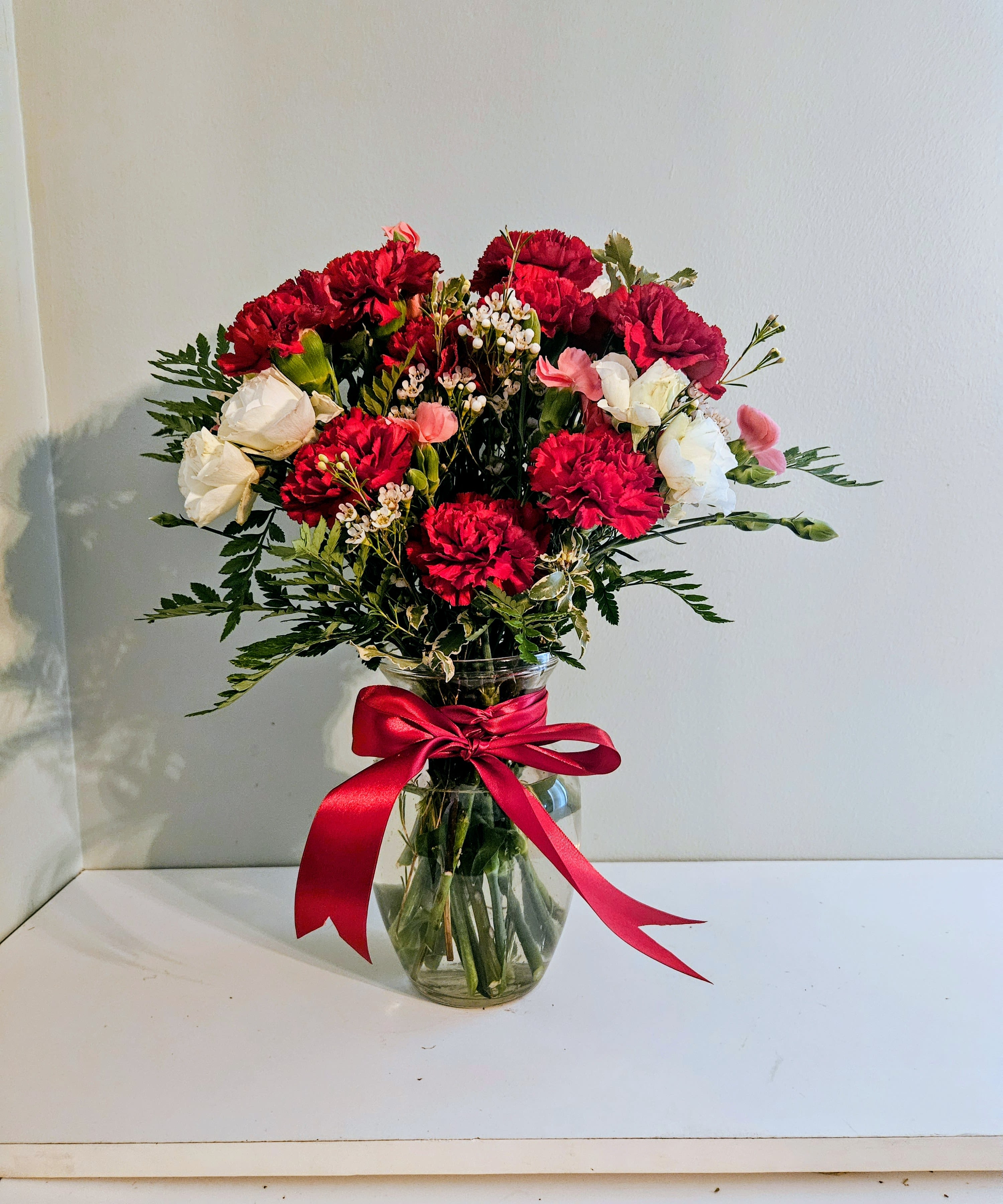 Consistent Love - A rounded cluster of red carnations with white blooms and soft pink accents creates a cheerful, romantic flower arrangement perfect for an anniversary, Valentine's Day, or any time you want to show steady affection. The compact, gift-ready shape reads classic and confident-the person you're thinking of will feel truly seen by this timeless mix of red, white, and pink florals.  The arrangement is gathered in a clear glass vase and finished with a wide red satin ribbon for a polished presentation. Small filler blossoms and airy greenery add texture and lift, giving the bouquet a full, layered look while keeping the focus on the rich red carnations and delicate white and pink flowers. Colors: red, white, pink, and green; flowers include carnations plus complementary mixed blooms.  Made to arrive ready to display, this arrangement suits everyday gifting as well as special romantic moments. Care is simple: keep fresh water in the vase and trim stems if you transfer them to another container; exact dimensions and flower counts are as shown in the product images.