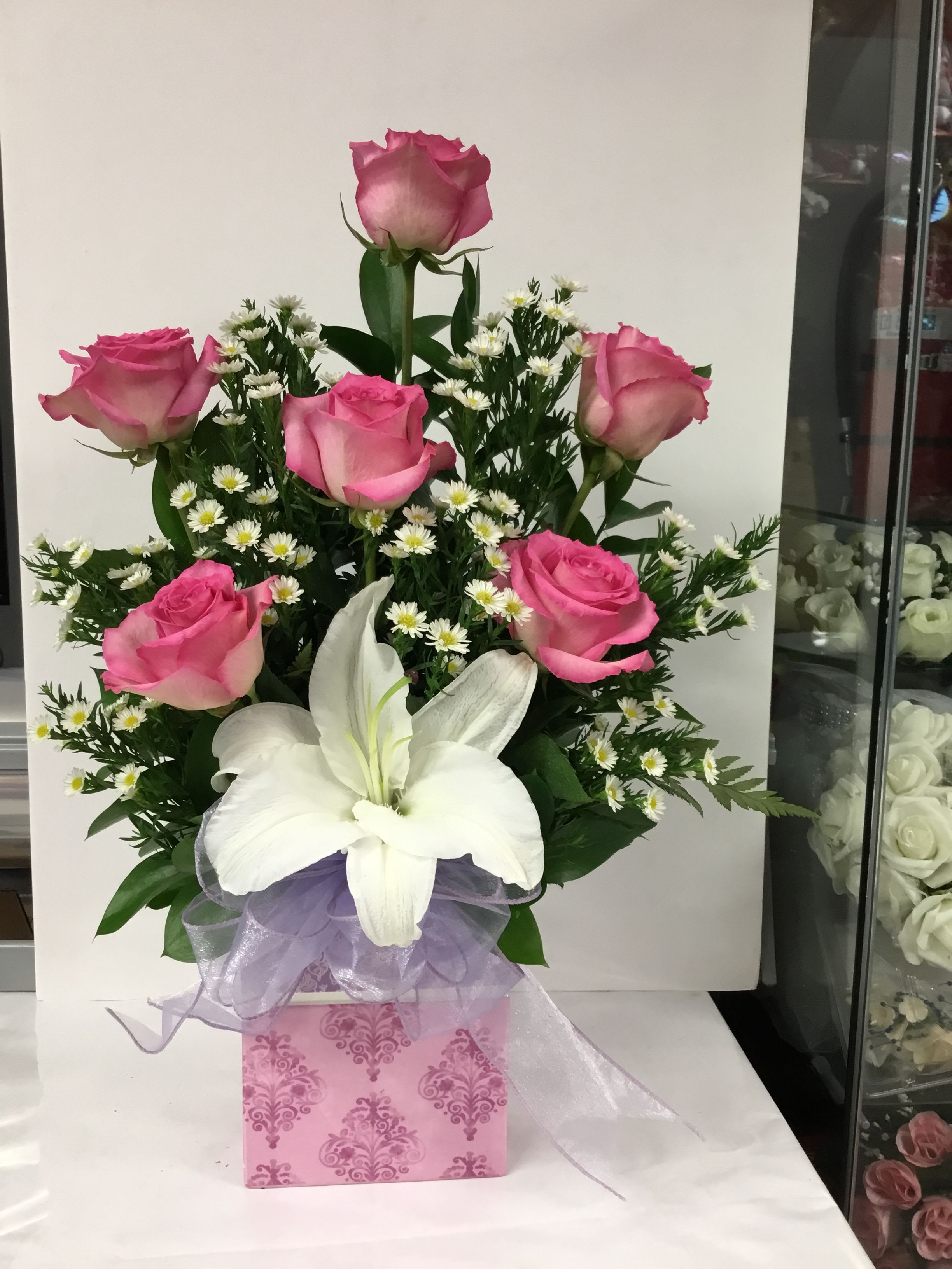 MOTHERS EMBRACE  - Soft pink roses clustered around a single dramatic white lily make this Mother's Day-ready flower arrangement an instantly romantic and comforting choice for birthdays, anniversaries, or a thoughtful "just because" surprise. The upright, full design puts the lily forward as a luminous focal point while pink roses and delicate white daisy-like fillers create a lush, tactile halo that reads feminine and heartfelt-the kind of bouquet that makes the person you're thinking of feel truly seen.  Finished in a pink patterned gift box with a lavender sheer ribbon, the arrangement mixes shades of pink, white, green foliage, and a touch of purple for gentle contrast. The roses form the main body and provide soft, velvety texture; the lilies add scent and visual drama; and the small daisy fillers lighten the composition with airy detail. Packaged ready to present, it's ideal for marking birthdays, anniversaries, moments of love and romance, or simply brightening someone's day-no special care beyond fresh water is needed to keep the blooms looking their best.