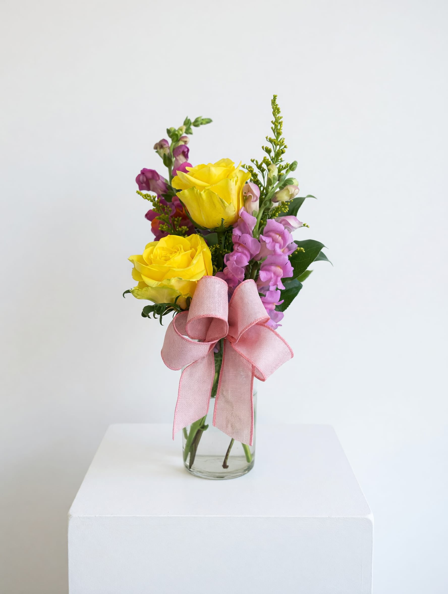 Sunshine and Love - Two bright yellow roses set the tone in this cheerful flower arrangement, paired with pink snapdragons and small yellow filler accents to create a fresh, light romantic feel-perfect as a birthday bouquet, a thank-you gesture, or a just-because surprise. The compact upright shape makes it an easy gift for someone you want to brighten today, the kind of arrangement that helps the recipient feel noticed and genuinely celebrated.  Arranged in a clear glass vase and finished with a wide pink ribbon, the mix of yellow, pink, green, and white reads sunny and uplifted without feeling overdone. The taller snapdragons add vertical texture against the rounded rose heads while the small fillers provide a soft, airy finish. Approximately sized for tabletops and desks, this gift-ready presentation needs only simple fresh water changes; trim stems and refresh the water every few days to keep blooms looking their best.