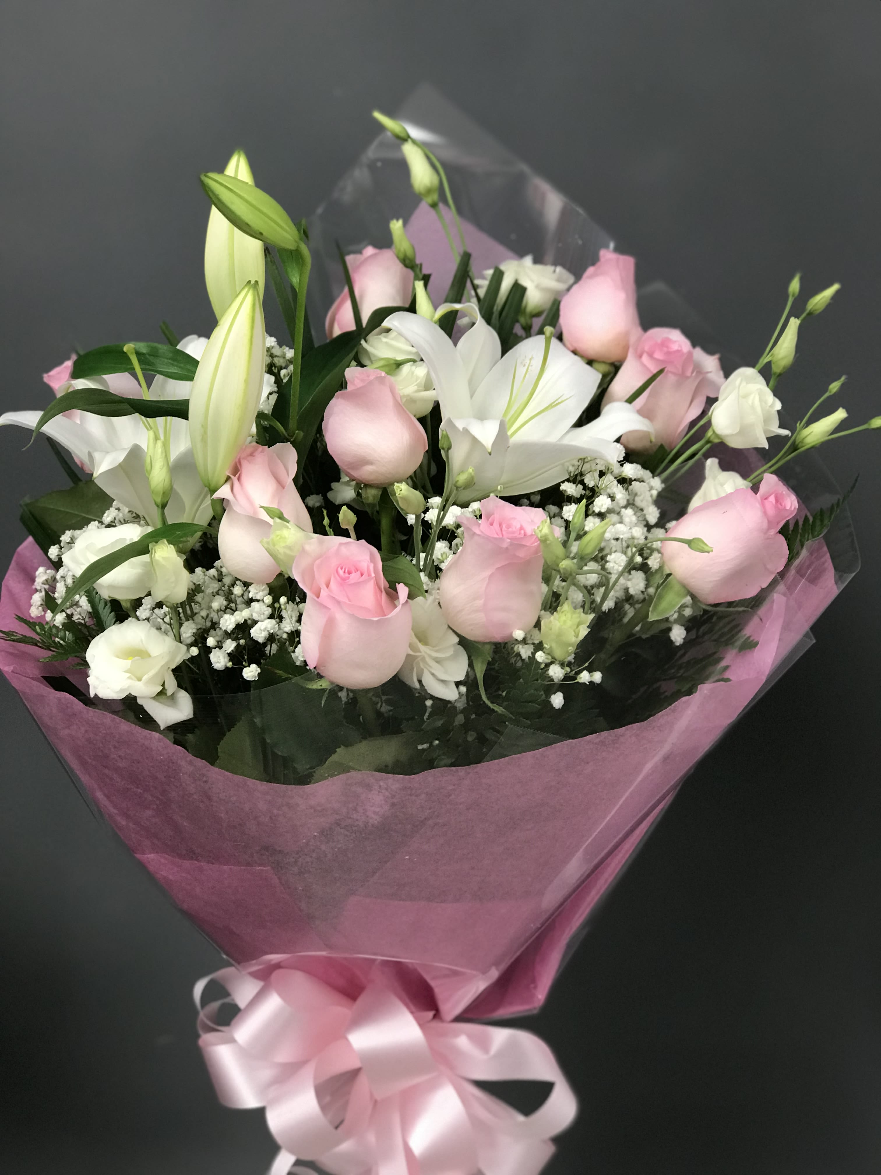 BLUSHING GLOW  - Pale pink roses paired with crisp white lilies create a soft, romantic flower arrangement perfect for an anniversary, birthday, or a love-and-romance gift that makes the person you're thinking of feel cherished. The rounded, hand-tied bouquet reads gentle and polished - the lilies add bright contrast and a touch of formality while the roses keep the mood intimate and tender.  This arrangement mixes blush roses, white lilies, and delicate white lisianthus and filler blooms for a textured, layered look in shades of pink, white, and fresh green. Blooms are gathered in a rounded hand-tied shape and wrapped in translucent pink paper with clear plastic wrap beneath, then finished with a pink satin ribbon for an elegant presentation that's ready to present.  Choose this bouquet when you want someone to feel noticed and celebrated - ideal for marking milestones, sending heartfelt apologies, or simply reminding a loved one they're adored. Follow the usual care for cut flowers: refresh water daily, trim stems at an angle, and keep away from direct heat to extend the life of the blooms.