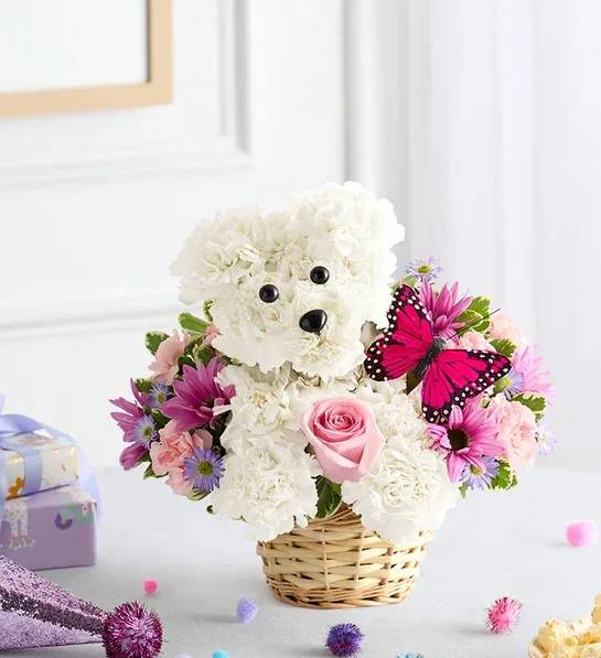 Precious Pup - One-sided, 3D arrangement with white carnations crafted in the shape of a dog complete with eyes and nose; surrounded by a mix of pink roses, light pink mini carnations and purple monte casino; accented with assorted greenery and butterfly pick