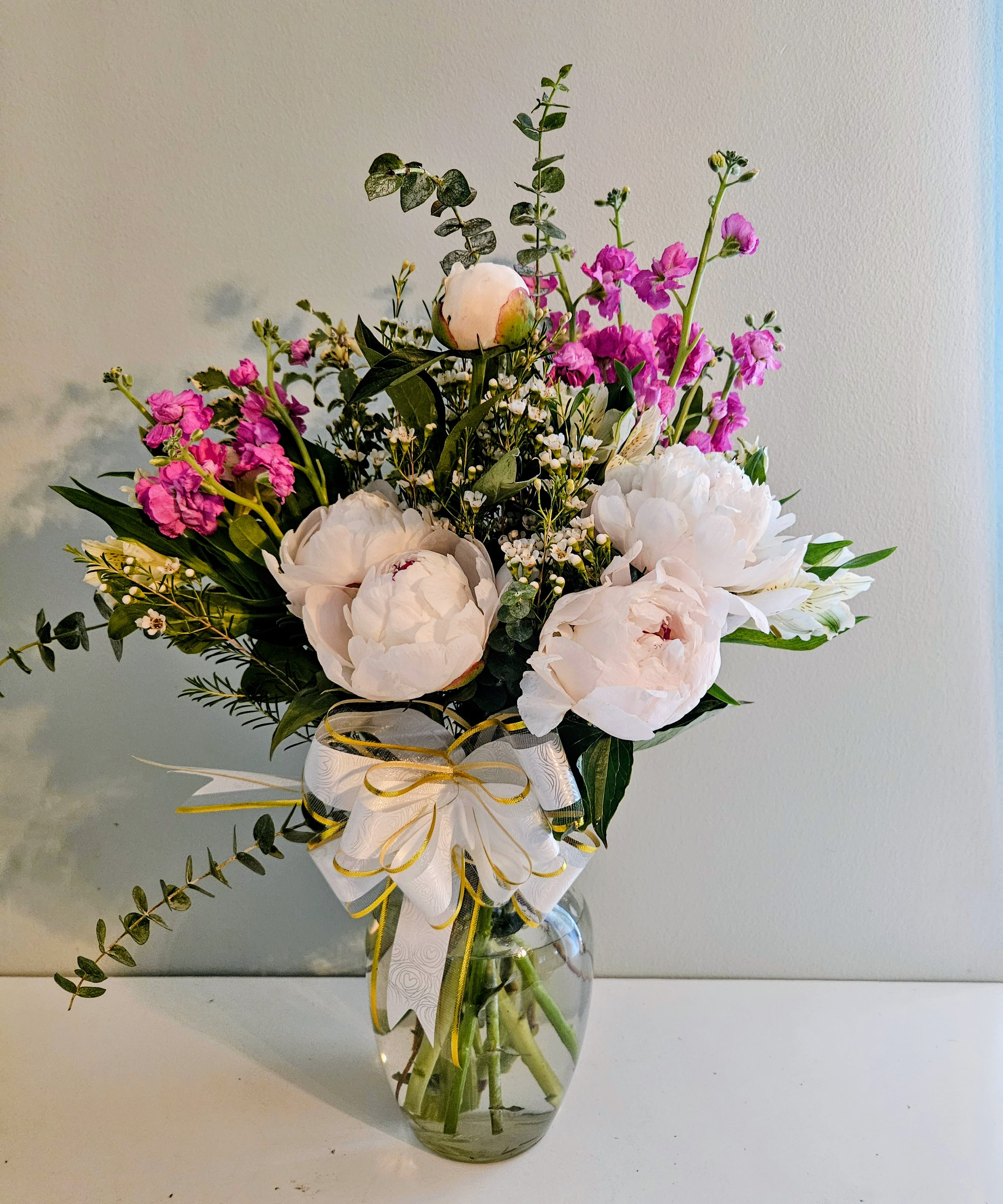 Peony Harmony - Big, cloud-like white peonies take center stage in this Peony Harmony flower arrangement, a fresh and romantic bouquet perfect for a birthday, thank-you gift, or spring celebration. The combination of soft white blooms with bright pink stock and mixed greenery creates an airy, joyful impression that will make the person you're thinking of feel cherished and celebrated.  This hand-tied arrangement is gathered in a clear glass vase and finished with a crisp white wrap and a yellow-edged decorative ribbon for a polished, present-ready look. Small white filler flowers add texture and fullness alongside the peonies and stock, while the mixed greens provide contrast and depth. The palette of white, pink and green reads gentle but vibrant-an elegant choice for everyday gifting or to mark a special moment.  Designed to arrive ready-to-display, Peony Harmony suits mothers, friends, or loved ones who appreciate soft, romantic florals. For best longevity, keep in fresh water and out of direct heat; replace vase water and recut stems every few days to help the peonies open fully and last longer.