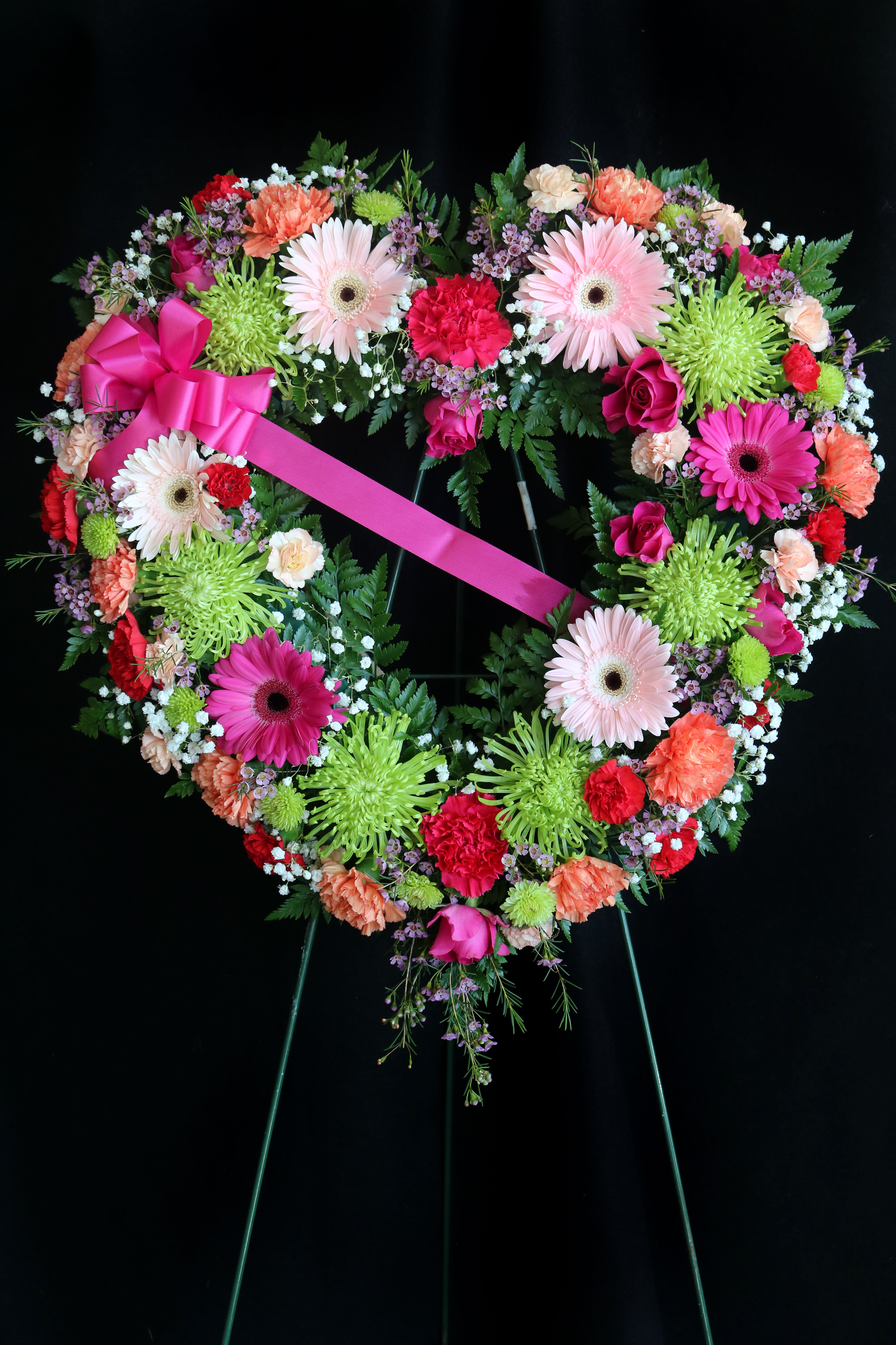 Heart-shaped floral wreath with a pink ribbon bow on a black stand