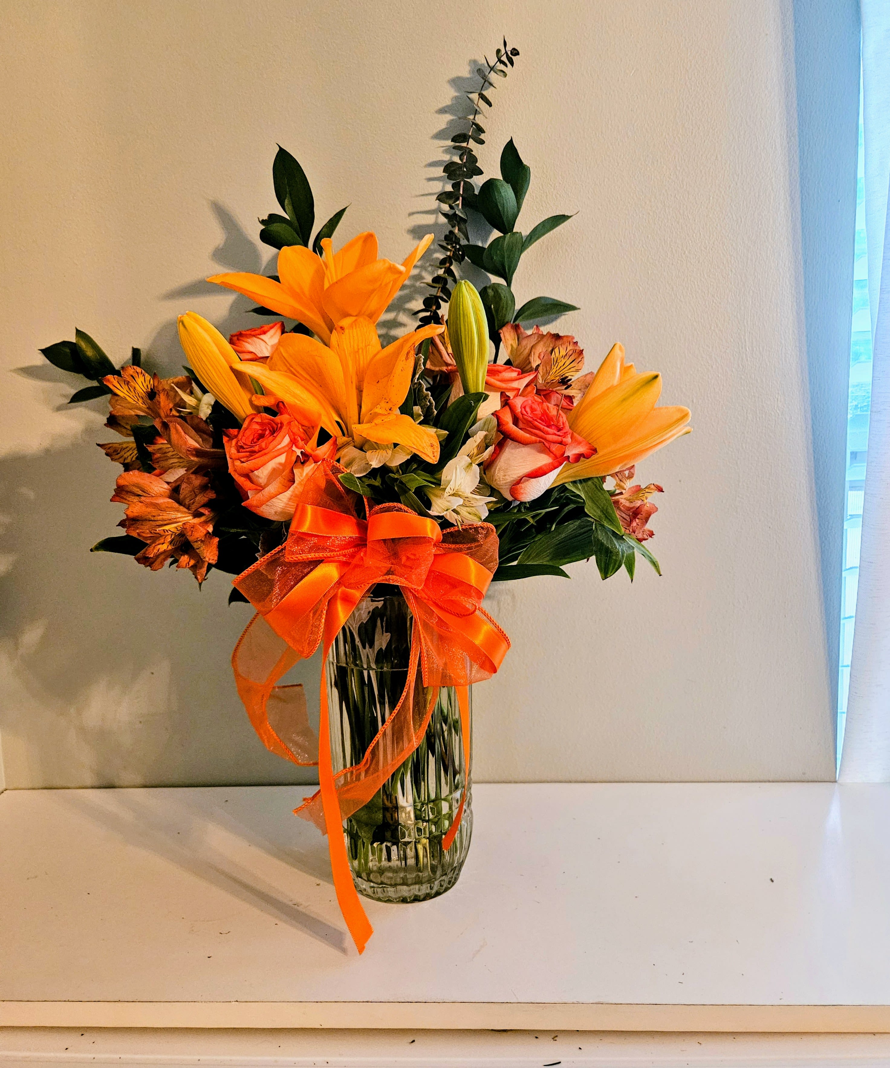 Passionate Love - Bright orange lilies take center stage in this Passionate Love flower arrangement, making it an ideal anniversary or celebratory bouquet that feels joyful and full of warmth. The mix of orange-pink roses and delicate white alstroemeria softens the palette, so the person you're sending it to will feel both noticed and genuinely celebrated-perfect for birthdays, congratulations, or "just because" moments.  Arranged in a clear ribbed glass vase and finished with a large sheer orange ribbon around the neck, the bouquet has a lively, energetic silhouette with fresh green foliage to balance the vivid colors. Lilies provide bold visual impact and a light, sweet scent, while roses add romantic depth and alstroemeria brings a cheerful, textured touch.  This design suits anyone who loves bright, uplifting color and wants a gift that reads as thoughtfully joyful. Vase included; treat with fresh water and trim stems every few days to keep blooms looking their best.