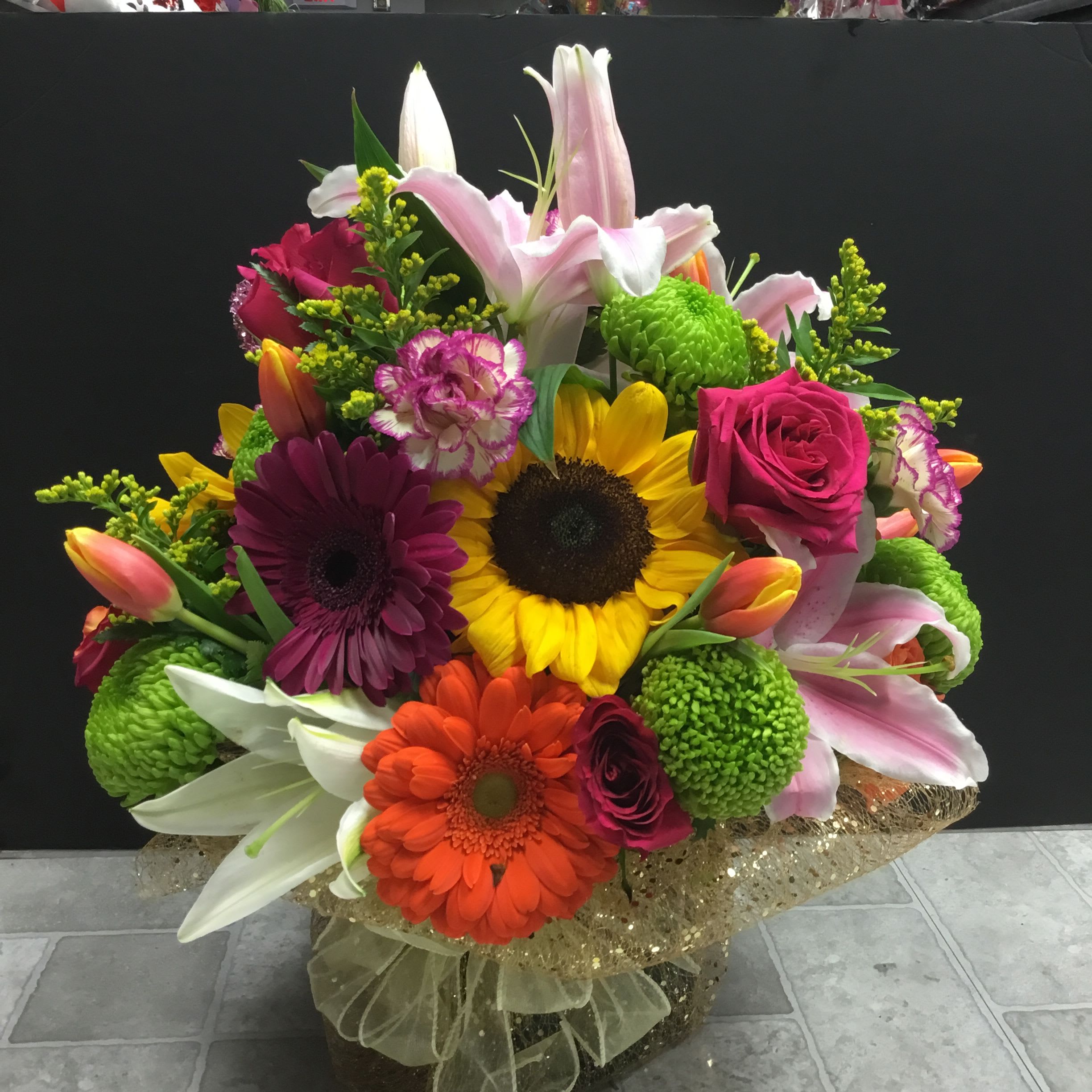 FLORAL FANTASY  - A bold, celebratory centerpiece that instantly brightens any birthday bouquet or congratulations arrangement-the dense, rounded shape of this flower arrangement catches the eye with a joyful mix of pink, yellow, orange and fresh green. Large lilies and vibrant gerbera daisies anchor the composition, giving the person you're celebrating a radiant focal point, while the overall palette reads cheerful and upbeat for anniversaries or just-because moments.  Roses, tulips, carnations and button chrysanthemums are layered around the focal blooms to create a full, textured look with lively depth and soft contrasts between petals. The arrangement sits in a clear vase and is finished at the base with a gold-toned mesh wrap for a slightly formal, festive touch that makes the gift feel special and intentional.  Perfect for someone who loves vivid color and generous blooms, this arrangement sends an unmistakable message of joy and celebration-she'll feel genuinely celebrated the moment it arrives. Delivered ready to display in the included clear vase; keep stems trimmed and water fresh to extend vase life.