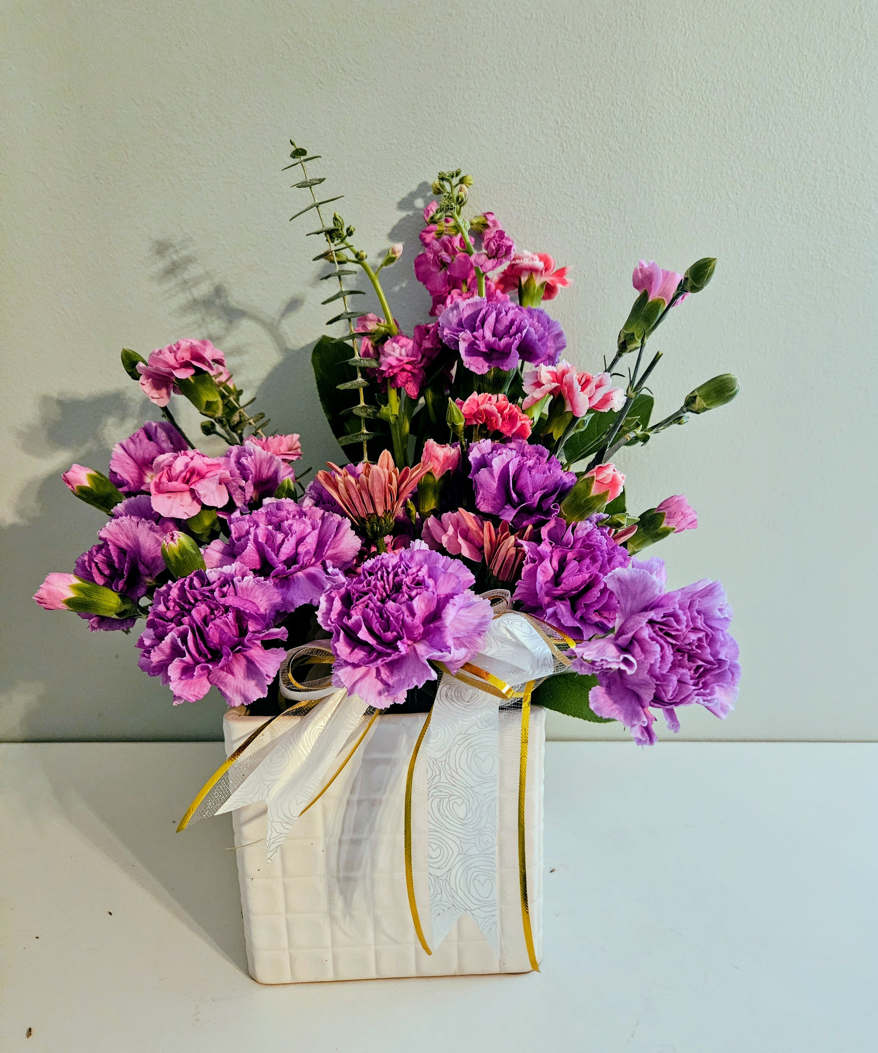 A Confident life - A confident burst of purple and pink carnations that brightens any birthday or thank-you moment-this flower arrangement is a cheerful, everyday gift ready to make someone feel truly celebrated. The compact bouquet reads slightly formal yet approachable, perfect for a spring celebration or to mark an encouraging milestone in a loved one's life.  Dense clusters of saturated purple and softer pink carnations form the main mass, with a few smaller accent blooms rising above for texture and movement. The stems sit neatly in a white square ceramic vase and are finished with layered white-and-gold ribbon for a polished presentation. The palette of purple, pink, and touches of white feels fresh and uplifting without being fussy.  Give this to someone who could use a confident, colorful reminder that they're appreciated-whether it's for a birthday, a job well done, or just because. The arrangement arrives ready to display in its vase; keep blooms in fresh water and out of direct heat to prolong their life.