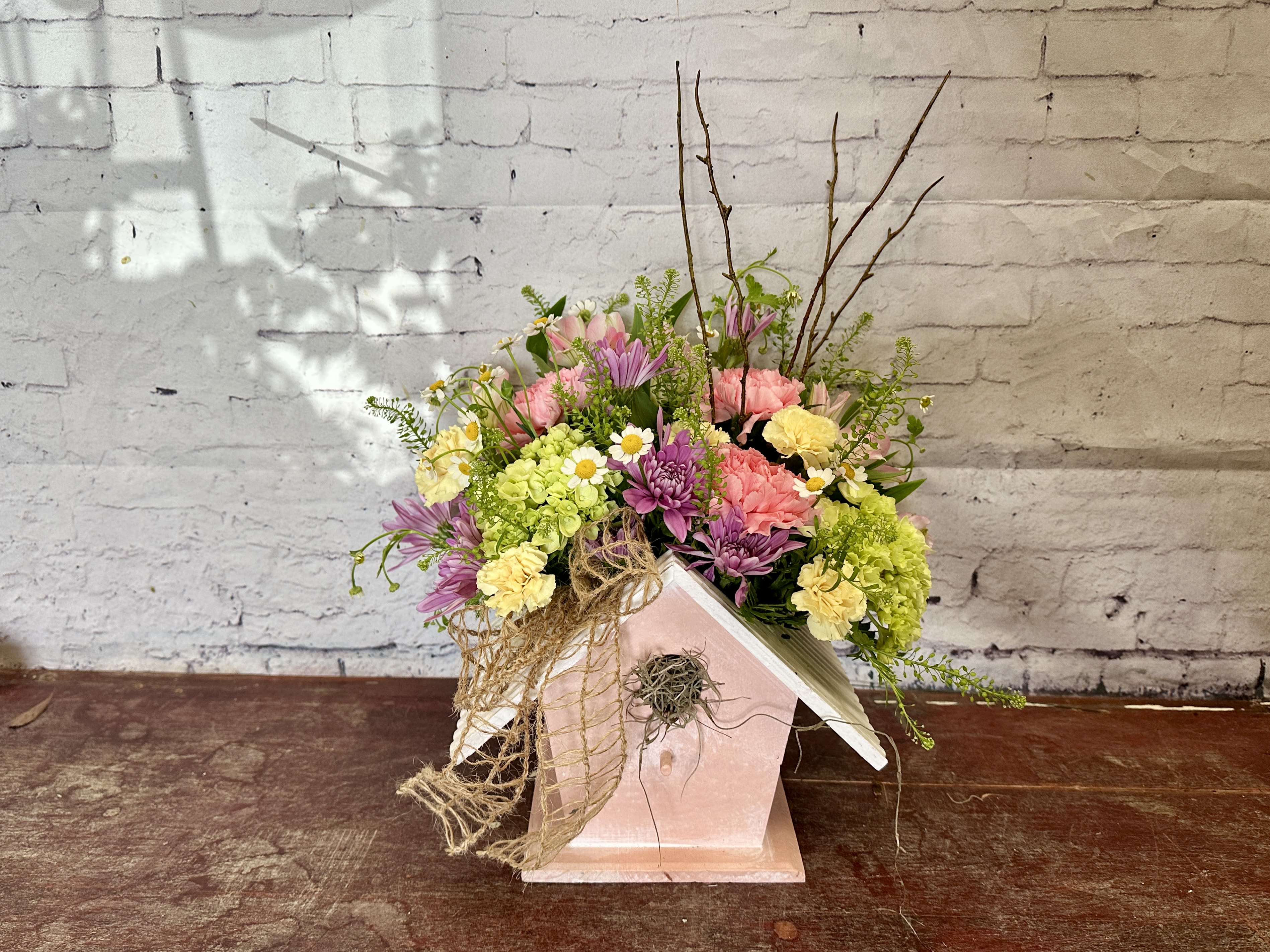 Birdhouse Blooms - Pastel pink, lavender, yellow &amp; white blooms in a birdhouse with carnations, daisies, hydrangea, twigs, moss and ribbon. Birdhouse color may vary. 