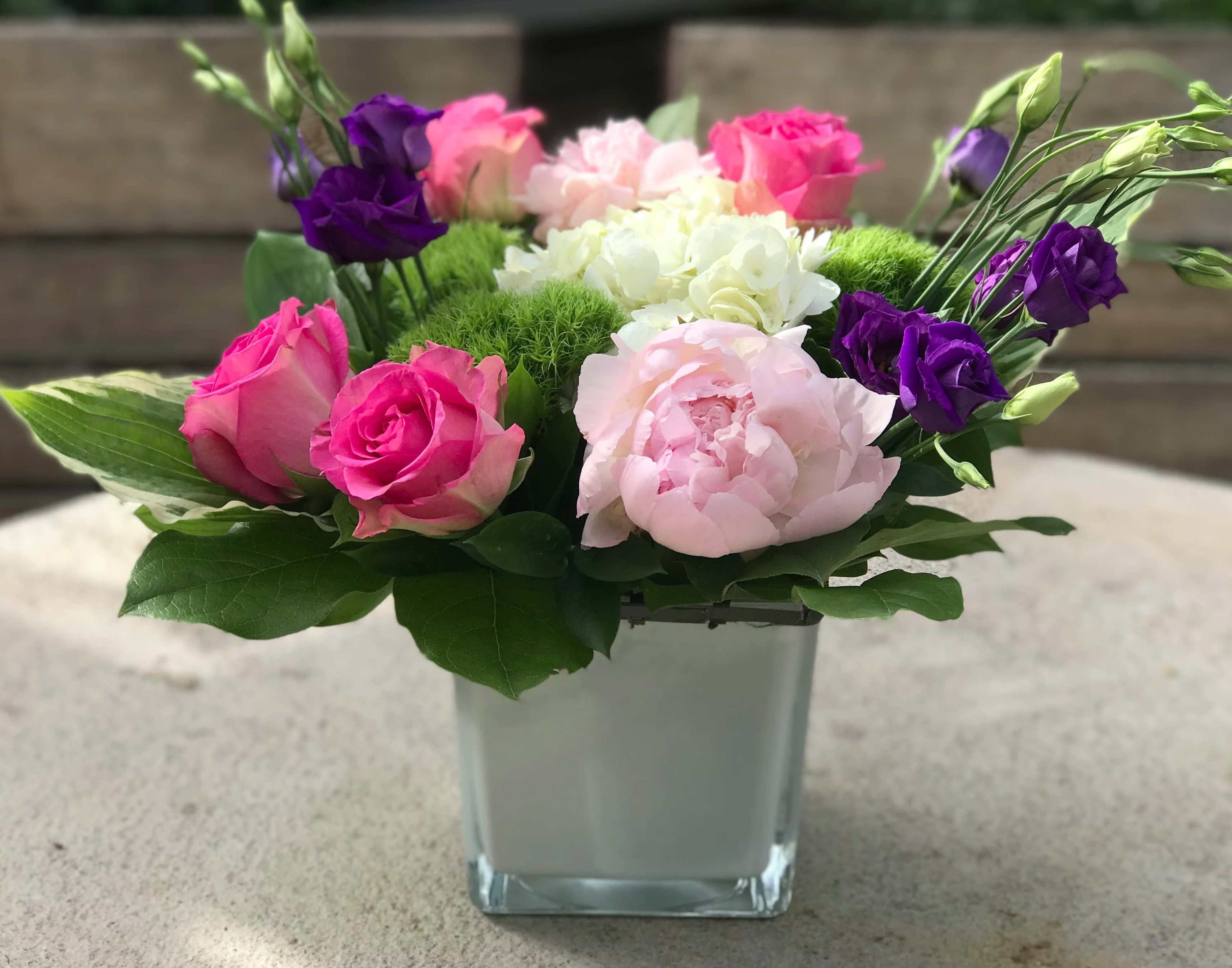 Low cube arrangement of pink roses, peonies, and purple lisianthus in a clear glass vase