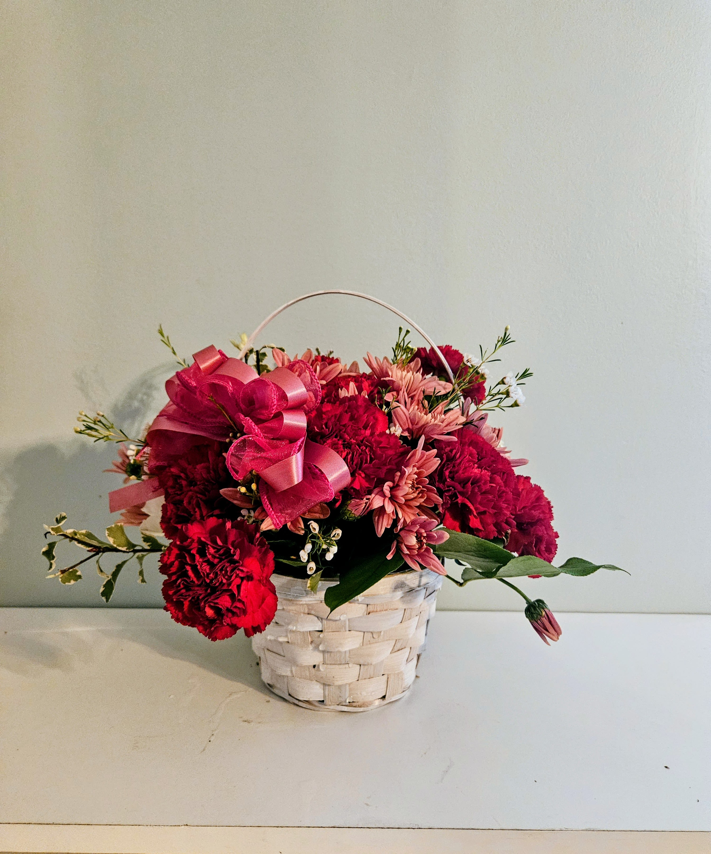 Sincerity Garden - A full, rounded cluster of pink and deep red blooms makes this flower arrangement feel like a sincere, heartwarming embrace - perfect as a birthday bouquet, anniversary gift, Mother's Day surprise, or a thoughtful thank-you arrangement. The mix of carnations and daisy-like chrysanthemums gives the bouquet a soft, romantic texture while the pop of red adds affectionate warmth that'll make the person you're thinking of feel genuinely seen.  The Sincerity Garden is arranged in a white woven basket with a comfortable handle and finished with a large pink ribbon bow for an extra touch of charm. Colors include soft pinks, rich reds, and crisp whites, with small white accent flowers adding delicate contrast. The composition is dense and rounded for immediate visual impact and a lush, garden-fresh look that stays appealing in any room.  Choose this arrangement when you want a cheerful, everyday gift that reads as both caring and elegant - ideal for moms, close friends, or a special someone marking a milestone. The basket presentation makes it display-ready with minimal fuss; simply place in a cool spot, refresh the water as needed, and enjoy the lasting color and texture of the carnations and chrysanthemums.