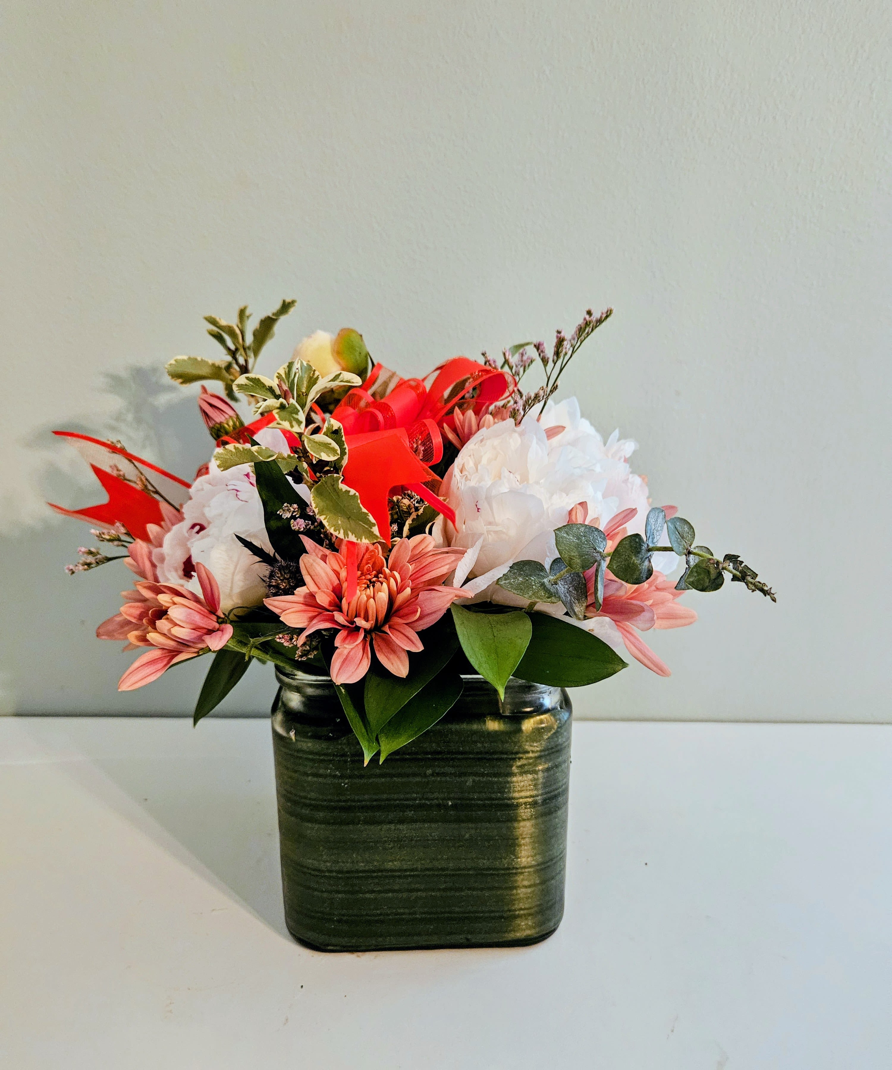 Wealth And Honor - A compact tableau of soft pink daisies, large white blooms, and a few red roses makes this flower arrangement an ideal birthday bouquet or everyday gift that reads both cheerful and gently romantic. The rounded composition sits in a dark green ceramic vase, tied with vivid red ribbon loops that lift the pastel palette and send a clear message of warmth and admiration - perfect for celebrating a loved one, saying congratulations, or brightening someone's day.  This arrangement features chrysanthemum-like pink daisy blooms, generous white blossoms, and additional mixed accents for texture, plus select red roses for contrast. The short, hand-arranged stems create a full, tabletop-friendly silhouette; the dark green ceramic vase and red ribbon give it a polished, finished look while keeping care simple. Designed to feel thoughtfully appointed without being fussy, it's the kind of gift that will make the person you're thinking of feel truly seen.