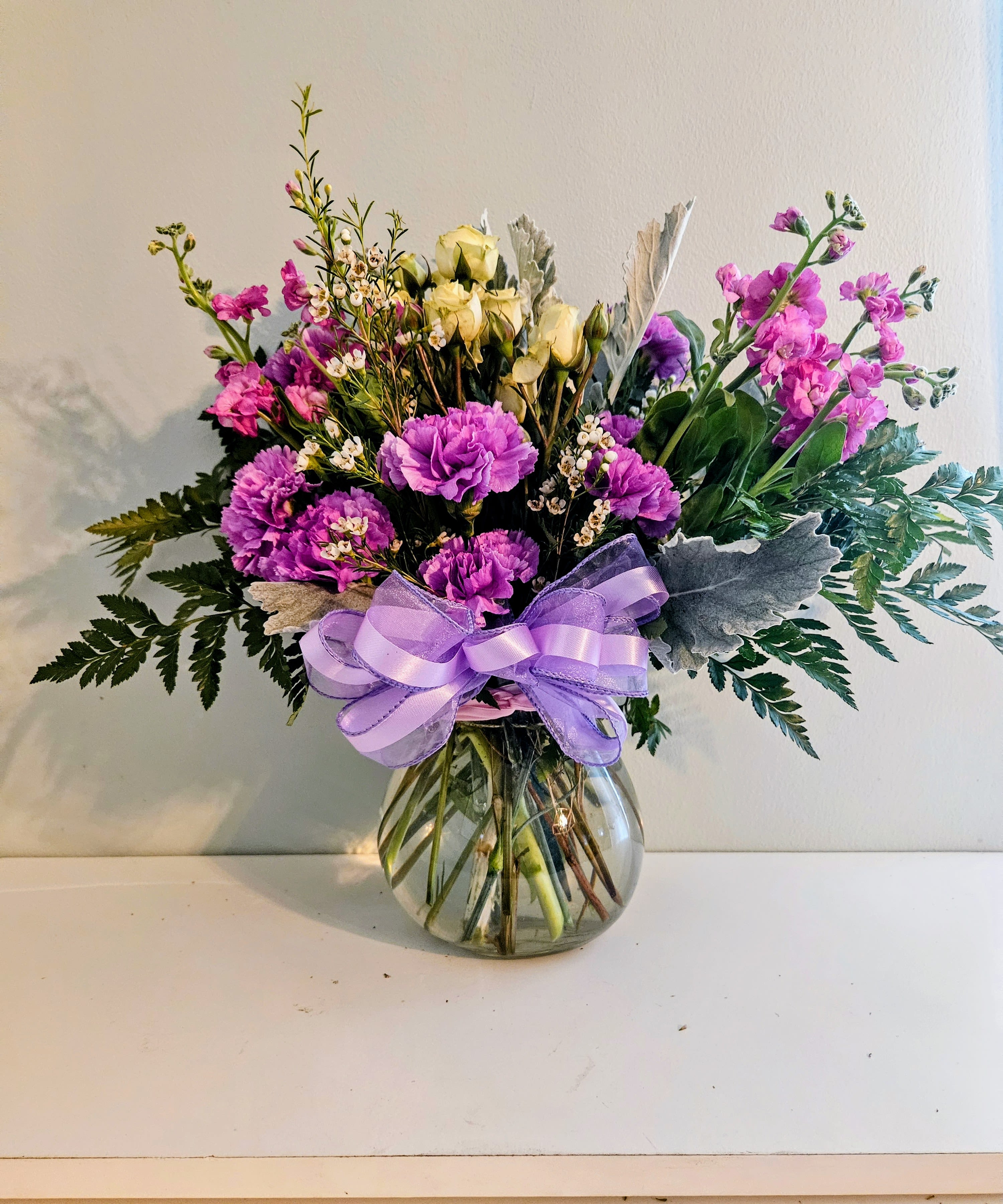 Purple Swing - Vivid purple carnations and soft pink stock-style blooms take center stage in this cheerful garden-style flower arrangement, perfect as a birthday bouquet, thank-you gift, or spring celebration centerpiece. The rounded composition feels slightly romantic and bright-designed to make the person you're thinking of feel noticed and celebrated the moment they see it.  Creamy pale roses nestle near the center, balanced by small white filler flowers and mixed green foliage for texture and contrast. Arranged in a clear glass vase and finished with a wide lavender ribbon and decorative silver foliage, the display reads lush and polished without being formal.  This is the kind of everyday gift that delights whether sent to a friend, mother, or colleague; it sends a warm, uplifting message of appreciation or congratulations. Standard vase size makes it easy to place on a dining table or desk; replace vase water every 2-3 days and trim stems slightly to help blooms last longer.
