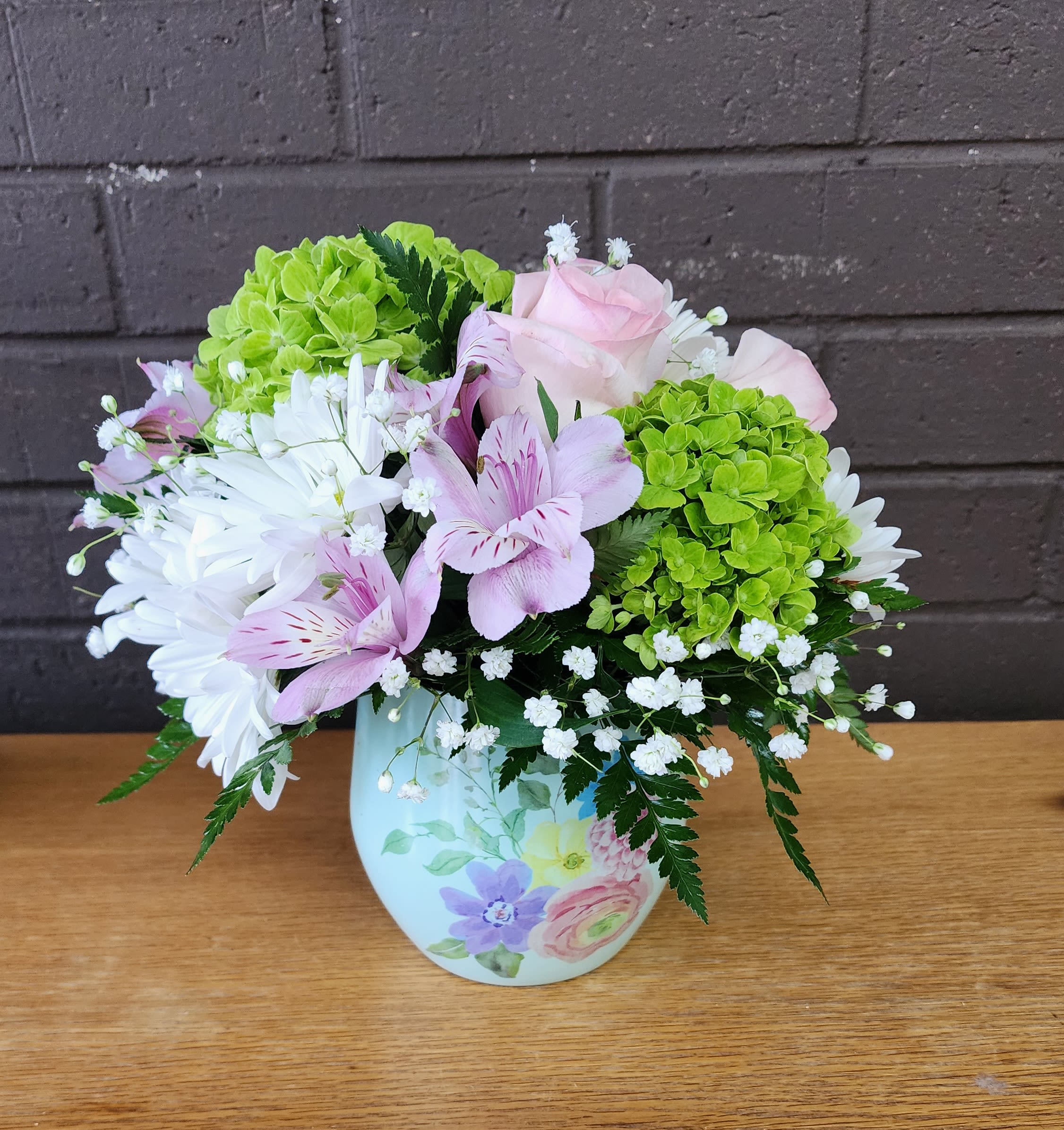Watercolor Garden - Soft green hydrangeas and pale pink roses create the first impression - a gentle, garden-fresh bouquet that reads like a watercolor painting. As a flower arrangement for an anniversary or a thoughtful get well gift, Watercolor Garden gives the person you're thinking of a calm, uplifting moment: the pale lavender alstroemeria and white daisy-like blooms mingle to brighten the room while sprays of baby's breath add an airy, comforting touch.  This full, rounded composition is arranged in a light-colored ceramic vase with painted floral decoration that reinforces the homey, hand-tended feel. Expect a mix of hydrangeas, roses, alstroemeria, daisies, and baby's breath in soft greens, pinks, whites, and purples - a palette that suits congratulations, Admin Professionals' Week, or simply saying "thinking of you." The vase makes it easy to display on a bedside table or desk; stems are arranged for immediate impact and simple care so the recipient can enjoy long-lasting color and gentle fragrance.
