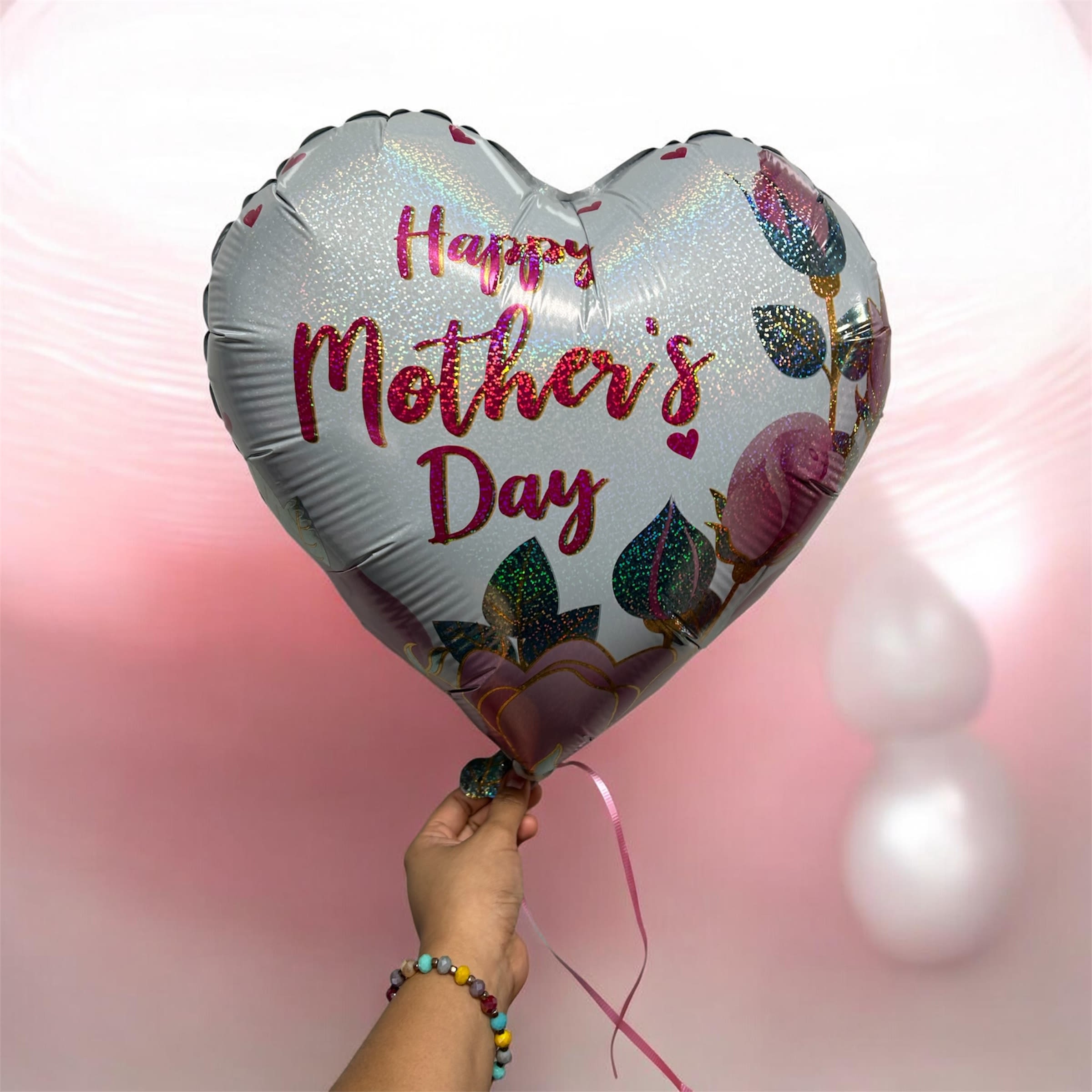 MOTHERS DAY  - A cheerful Mother's Day flower arrangement that pairs soft pinks, whites, and lavenders with a shimmering heart-shaped foil balloon-perfect for celebrating Mom, a grandmother, or any special woman who deserves to feel cherished. The silver-white balloon features glittery pink "Happy Mother's Day" lettering and small floral and heart accents, adding a festive, feminine touch that makes the whole gift feel thoughtful and joyful.  The bouquet combines delicate pink and white blooms with touches of purple for depth and contrast, tied with a pink ribbon for a polished, gift-ready look. The included heart balloon floats above the arrangement, creating instant visual impact and conveying warmth and appreciation. Ideal for Mother's Day, birthdays, or a simple "thank you" surprise, this arrangement arrives ready to display and delight the person you're celebrating.