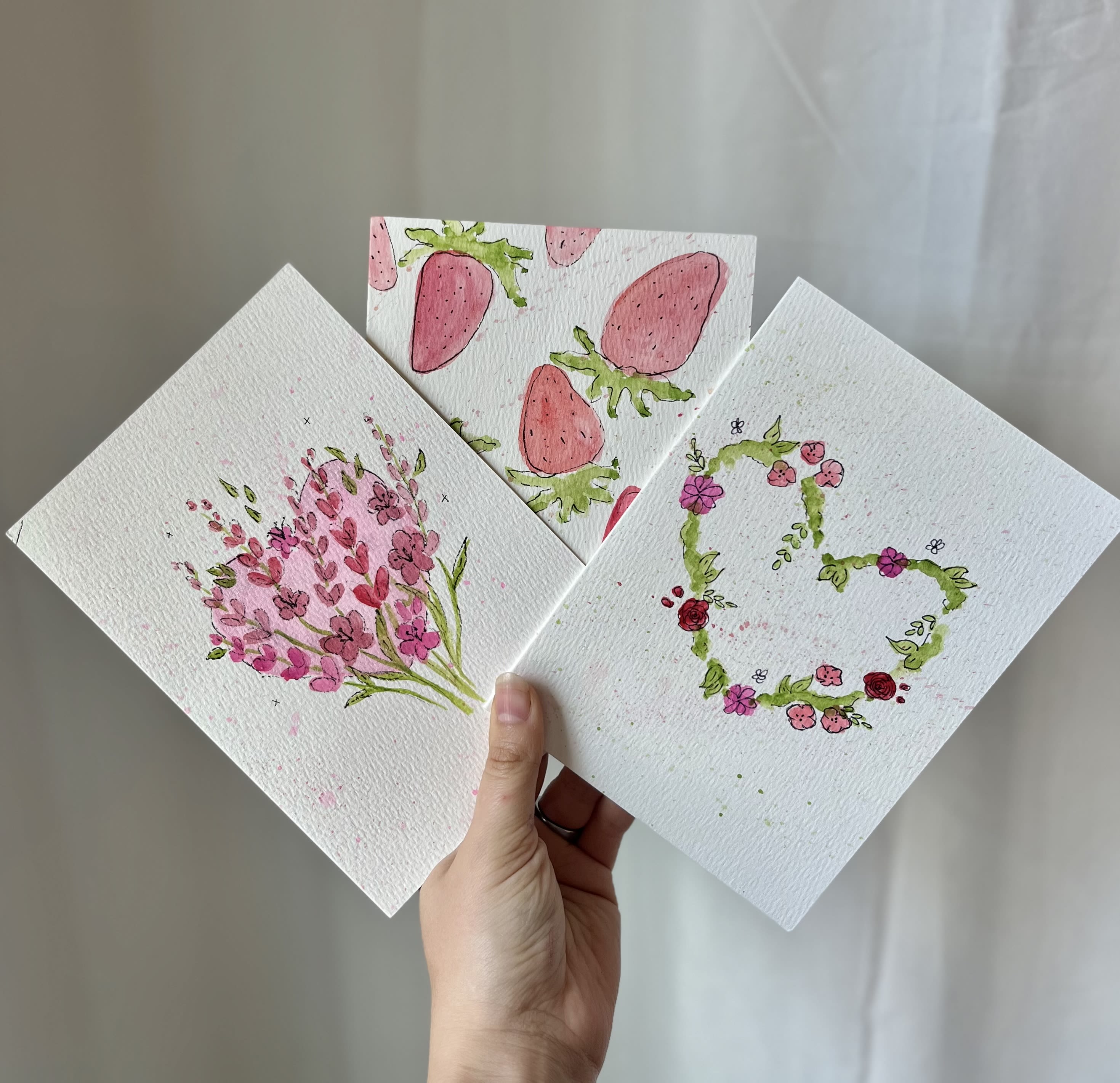 Hand holding three floral watercolor cards with pink designs