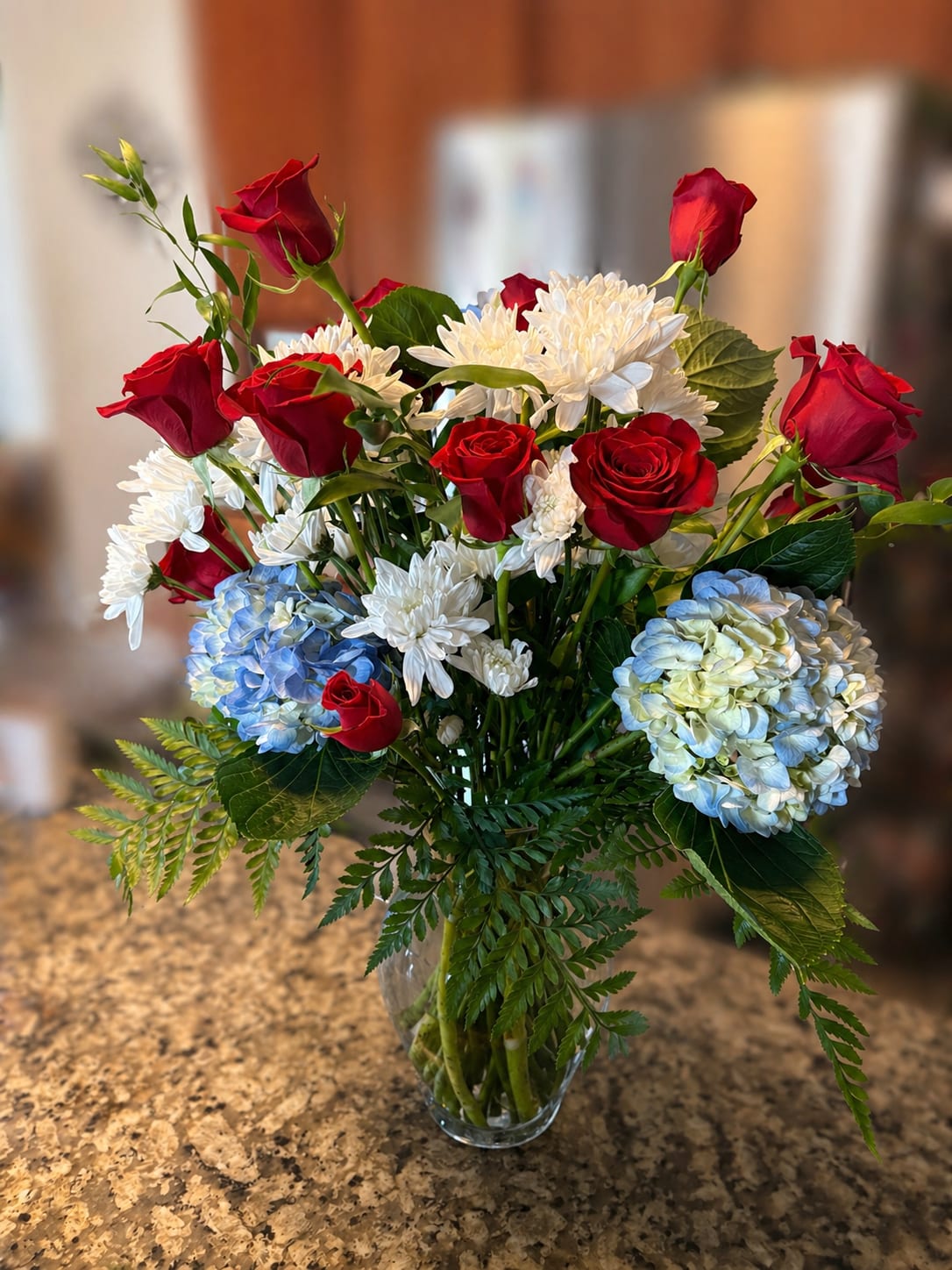 Red, White and Blue - A timeless blend of romance and calm, the Red, White and Blue arrangement pairs velvety red roses with soft white blooms and delicate blue hydrangeas. Accented with lush greenery, this arrangement strikes a perfect balance between bold passion and gentle elegance. Ideal for heartfelt moments, celebrations, or simply brightening a space, it brings warmth, beauty, and a touch of tranquility wherever it’s placed.