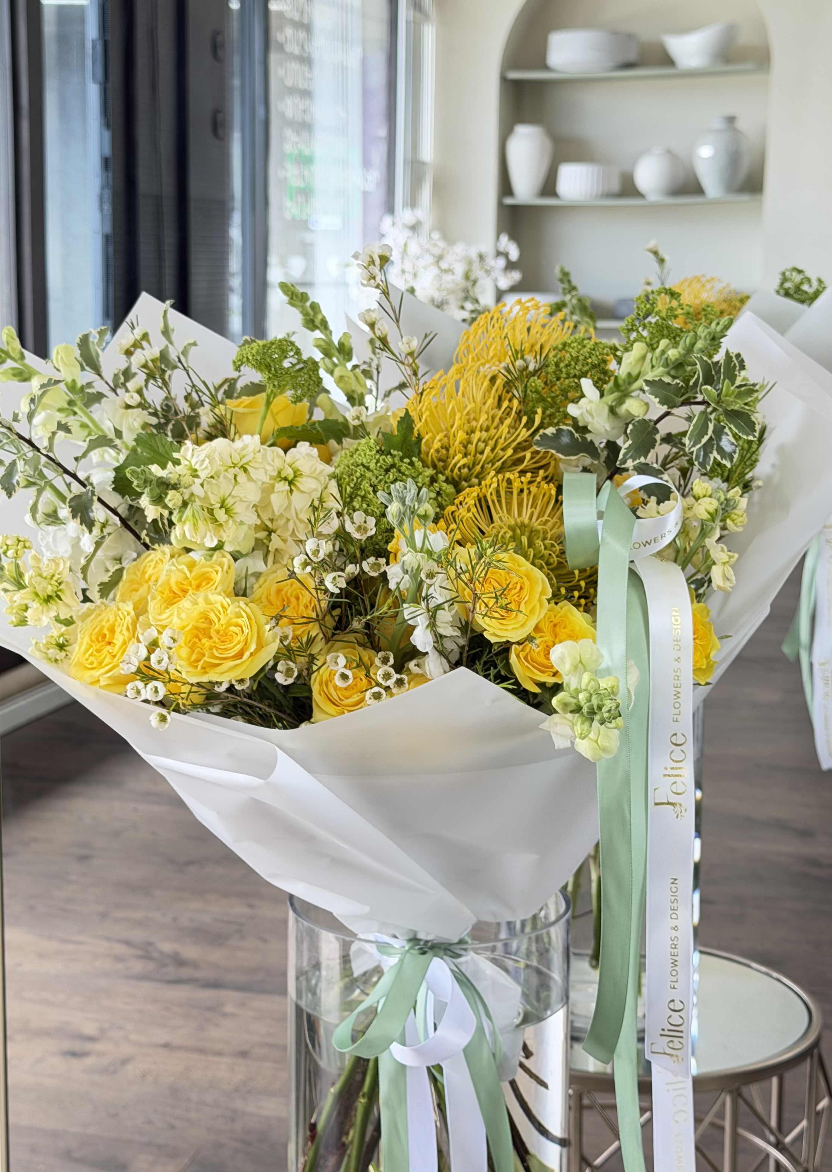 Golden Garden Luxury Bouquet - A vibrant and luxurious bouquet designed in fresh yellow, soft green, and creamy white tones, featuring premium garden roses, pincushion protea, hydrangeas, and seasonal blooms. This arrangement brings a bright and uplifting garden-style feel, combining texture, movement, and natural elegance. The golden palette creates a joyful and sophisticated look, perfect for making a statement. Carefully wrapped in signature white paper and finished with elegant ribbon detailing, this bouquet reflects the refined style of Felice Flowers &amp; Design. Perfect for Birthday, Congratulations, Get Well, New Baby, Thank You, and Just Because. Please note: due to seasonal availability, some flowers may be substituted. However, the overall color palette, bouquet size, and premium garden style will remain consistent with the photo.