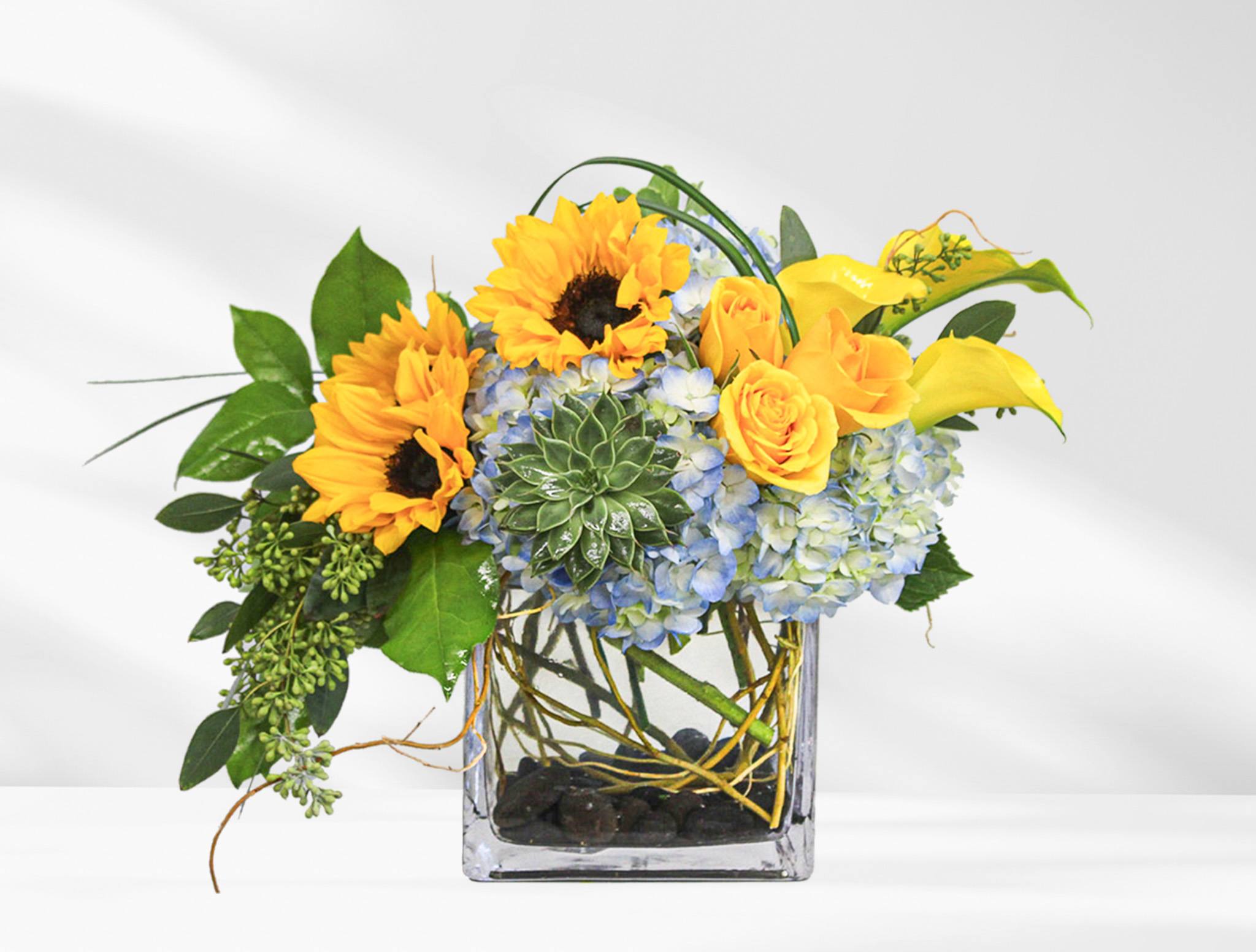 Sunflowers, yellow roses, blue hydrangeas, and calla lilies in a square glass vase