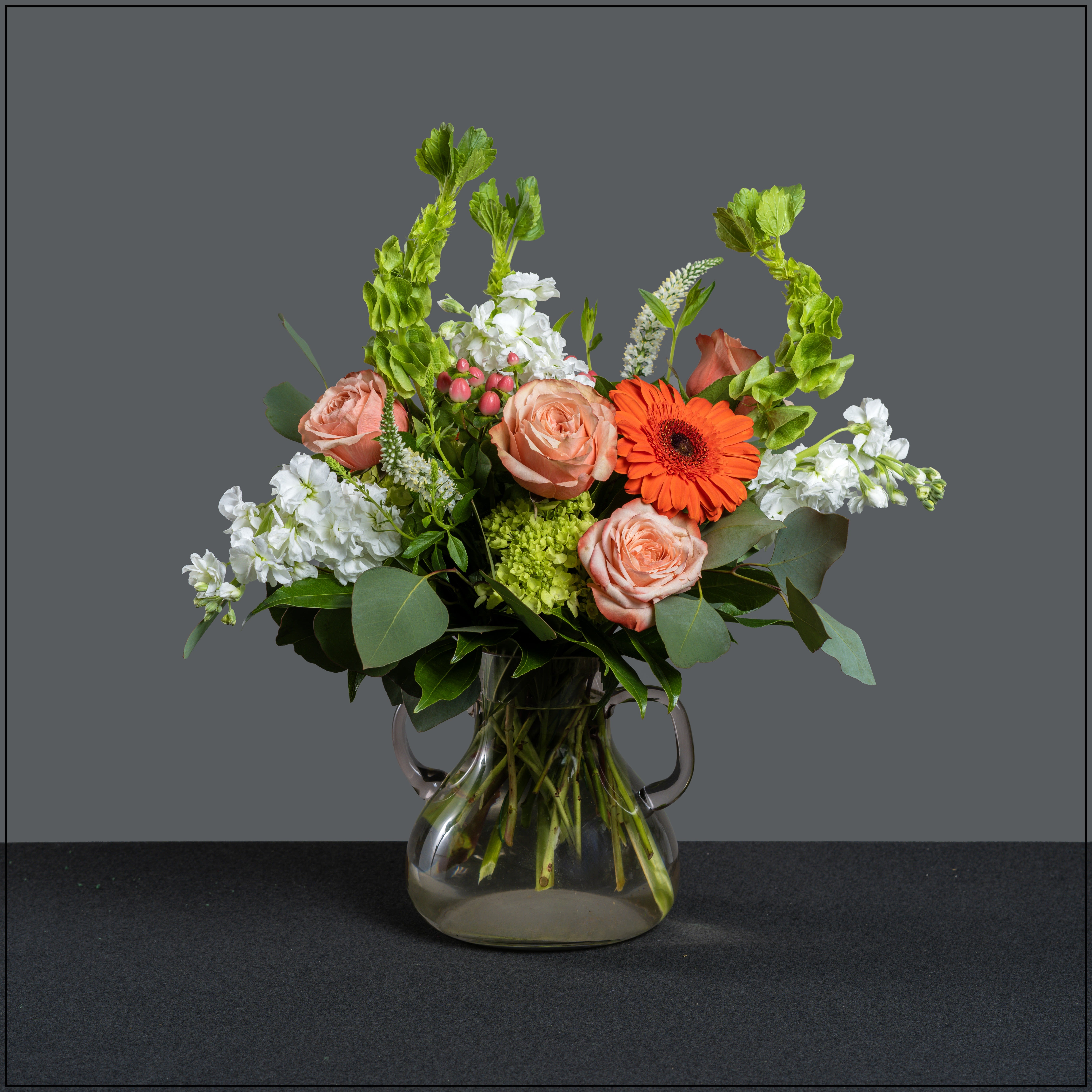 Beckett - Gorgeous glass vase with handles, filled with gerbera, roses, hydrangea, bells of Ireland, stock, veronica, and hypericum berries. Approximate size 16" tall and 20" wide. 