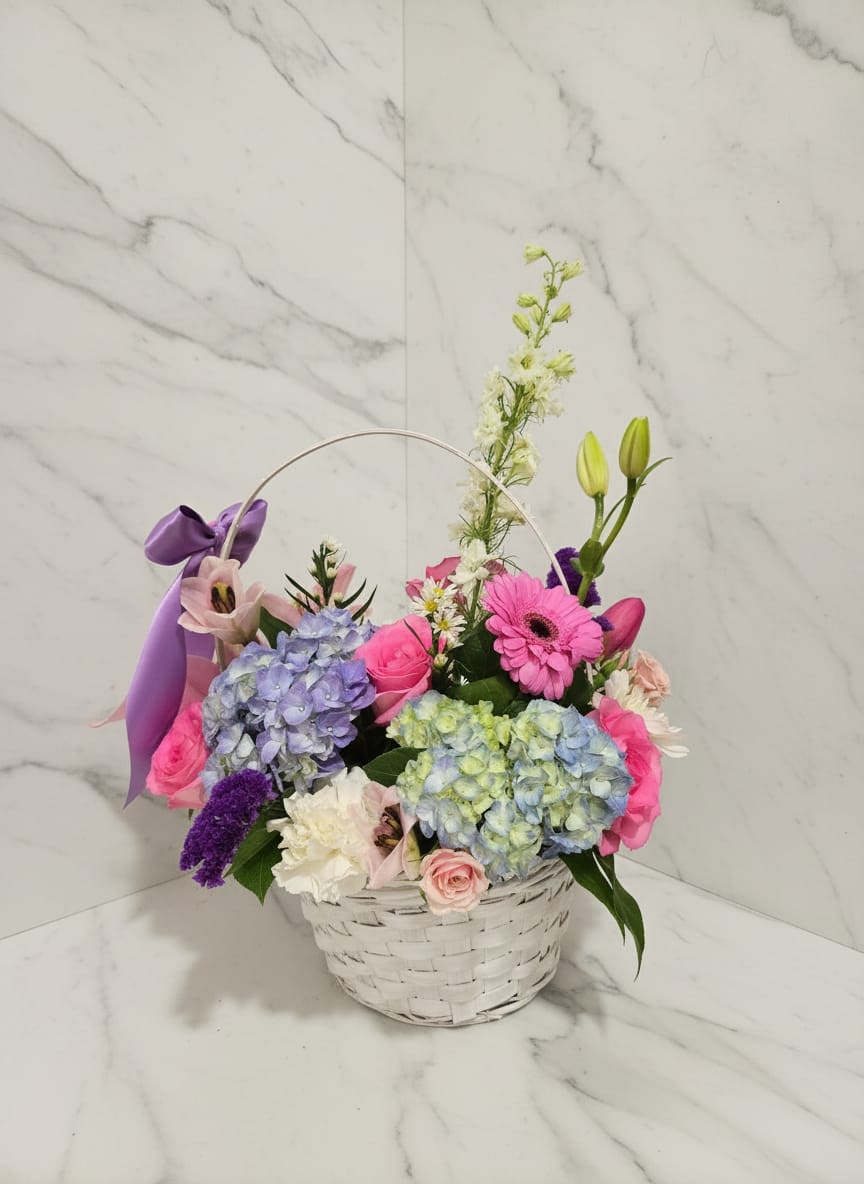 Pastel Picnic - Este arreglo hecho a mano es una clase magistral en textura y color, combinando hortensias suaves y etéreas con la presencia audaz y alegre de las margaritas Gerbera de color rosa intenso. Presentado en una clásica cesta de mimbre blanca y acabado con un lujoso lazo de satén lila, es un regalo que se siente tanto atemporal como fresco.