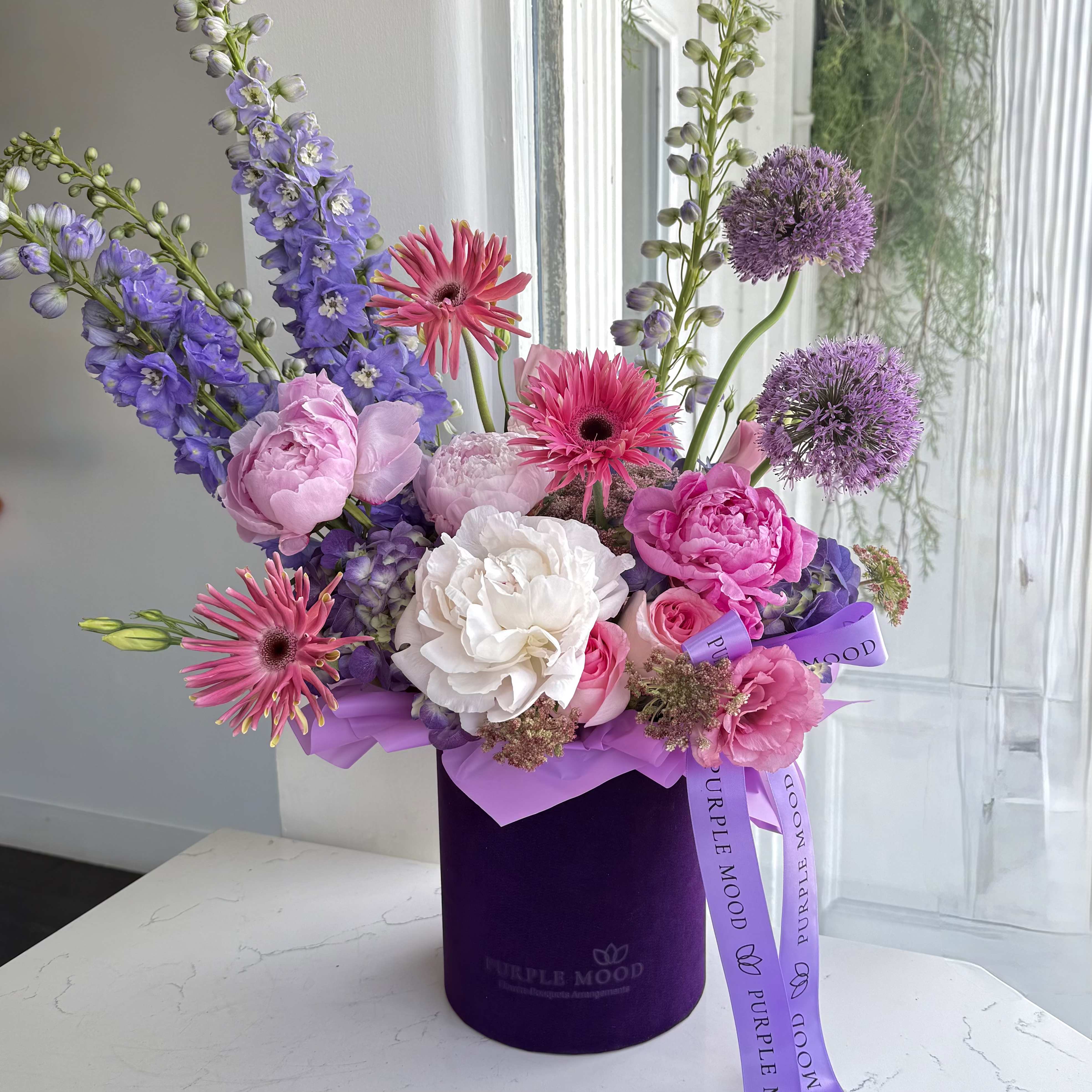 Purple Mood Box 360° - A lush, all-around floral arrangement designed to be enjoyed from every angle. This open-style box has no lid, allowing the blooms to flow freely in a full 360° display — perfect as a statement centerpiece.  Flowers are arranged in floral foam inside the box, so no vase is needed.