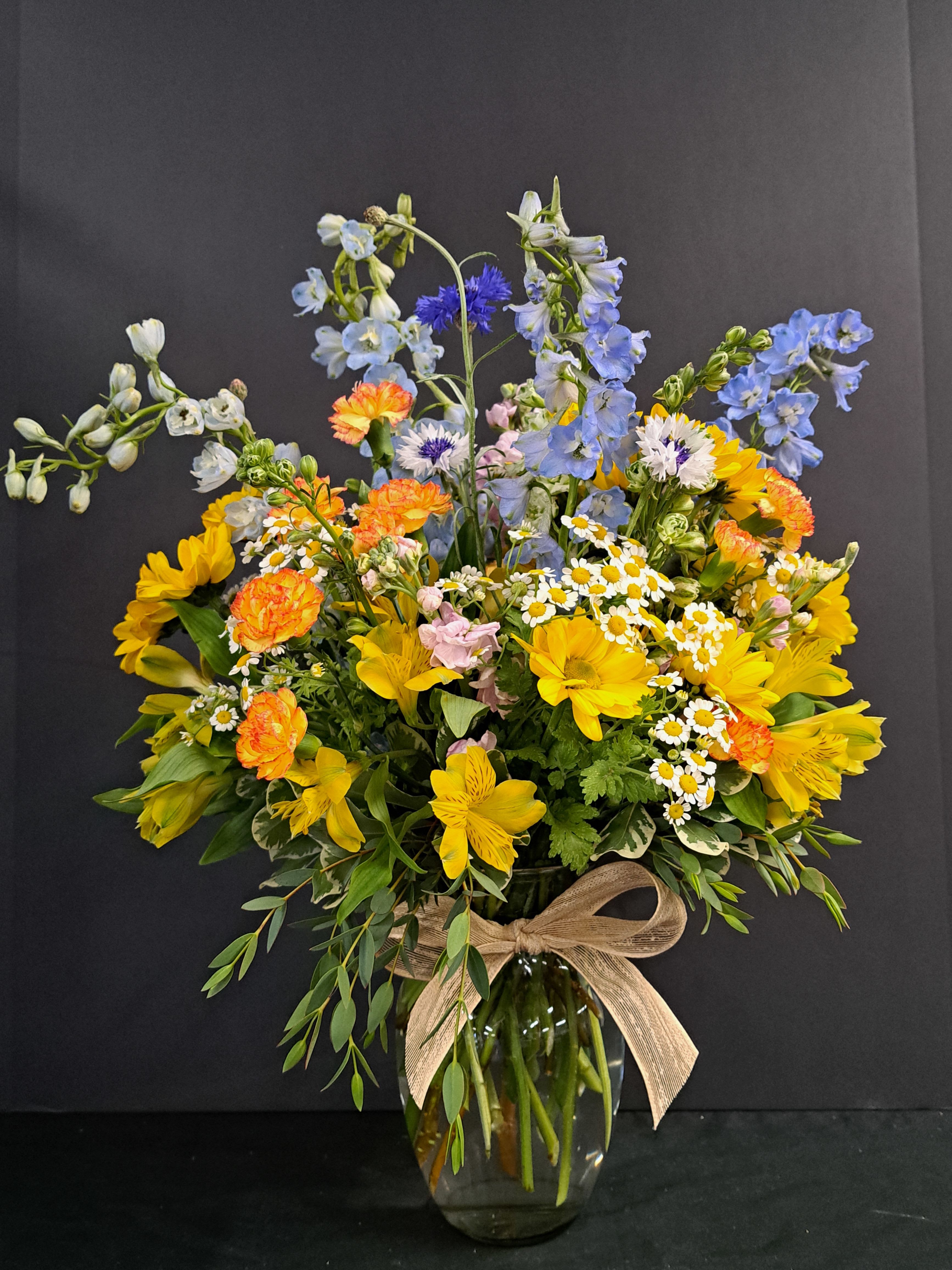 Mixed bouquet of yellow, blue, and orange flowers in a glass vase with a ribbon