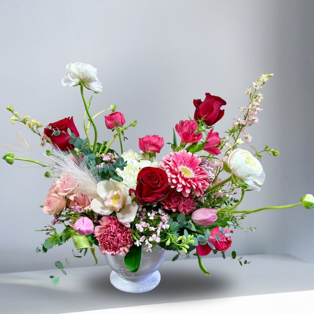 Blossoming Love - Recipe: This modern, garden look has seasonal ranuculus, red and hot pink roses, hydrangae, tulips, orchids, larkspur, orchid, carnations and more in a white vase. Seasonal: Seasonal Seasonal: All year round Orientation: Front Facing View Photo shown: Standard 