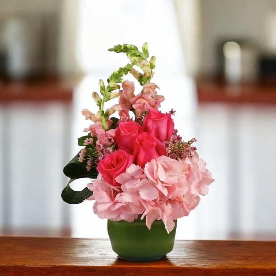 Majestic MOM - Bright pink roses paired with pale pink hydrangeas create a joyful, feminine birthday bouquet or anniversary arrangement that reads as both celebratory and thoughtful. The rounded clear cylinder vase lined with a ti leaf brings a fresh, earthy contrast that makes the pinks pop, while tall snapdragon stems add vertical interest so the recipient feels noticed and celebrated the moment they see it.  This design features clusters of soft, full hydrangea blooms nestled against vivid roses for a lush, textural look; snapdragons lift the composition and add graceful height. Housed in a compact clear cylinder lined with a ti leaf, the arrangement translates well as a centerpiece or a bedside pick-me-up and fits easily on a dining table or mantel.  Perfect for Mother's Day, a thank-you gift, or simply to brighten someone's day, this bouquet signals affection and cheer without being over the top. Arrives ready to display in its ceramic container; change the water every few days and trim stems if you transfer to a different vase to extend freshness.