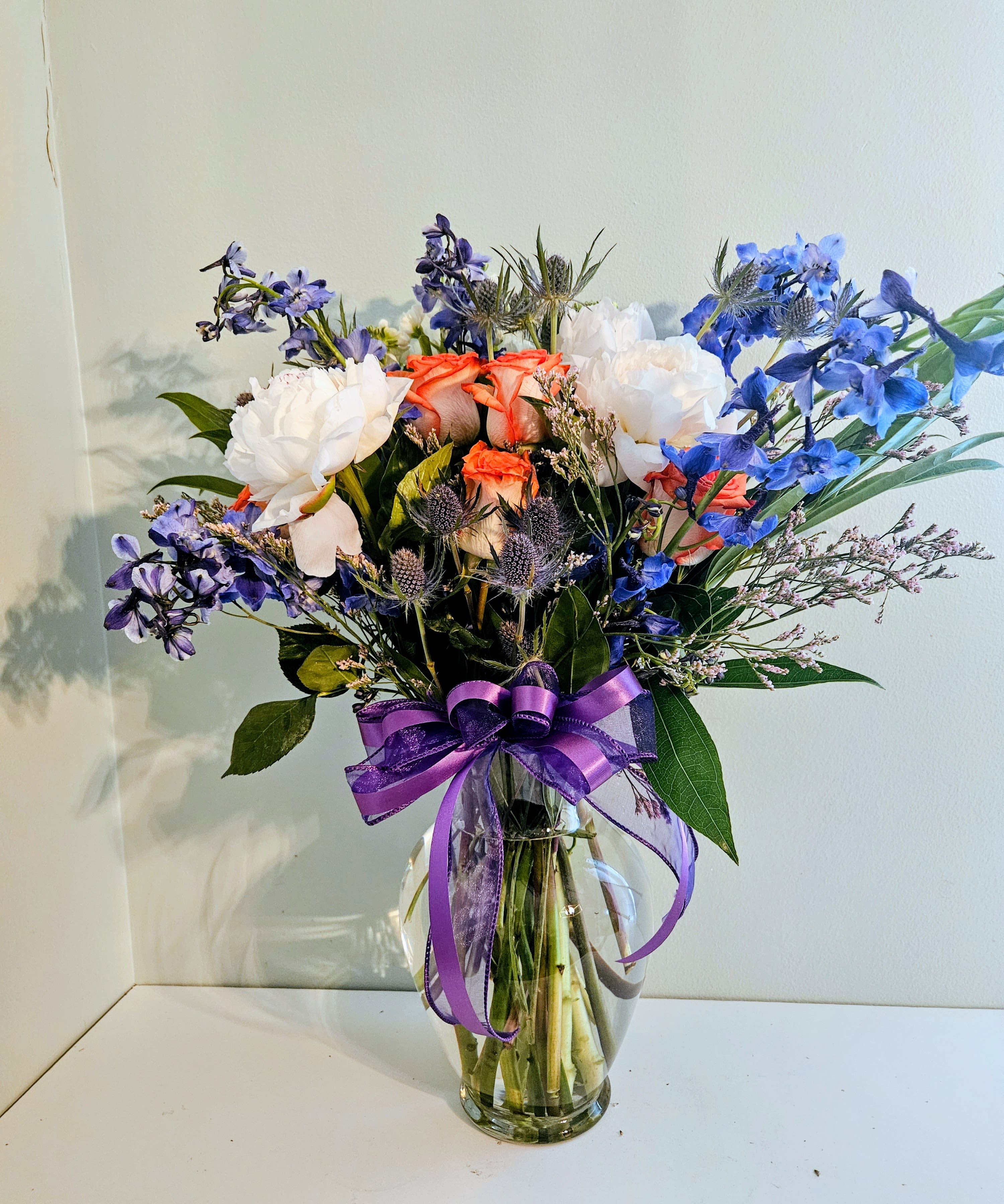 Elegance Life - A loose, airy bouquet where vivid blue delphinium-like spikes meet coral and white roses to create a fresh garden feel - perfect as a birthday bouquet, thank-you arrangement, or general celebration flower arrangement. The mix of cool blues, soft whites, and warm coral-pink gives the recipient an uplifting, slightly romantic impression that feels hand-picked rather than overly formal.  Arranged in a clear glass vase and finished with a wide purple ribbon at the neck, this composition balances structure and movement: tall blue verticals, rounded rose heads, and light filler blooms for texture. Colors in the bouquet read blue, white, pink, and purple; flowers include roses and delphinium-style blooms alongside mixed bouquet accents for variety and depth.  Send this when you want someone to feel noticed and celebrated - a thoughtful everyday gift that brightens a kitchen table or office desk. Vase-ready on delivery; trim stems and refresh water every 2-3 days to keep blooms looking their best.