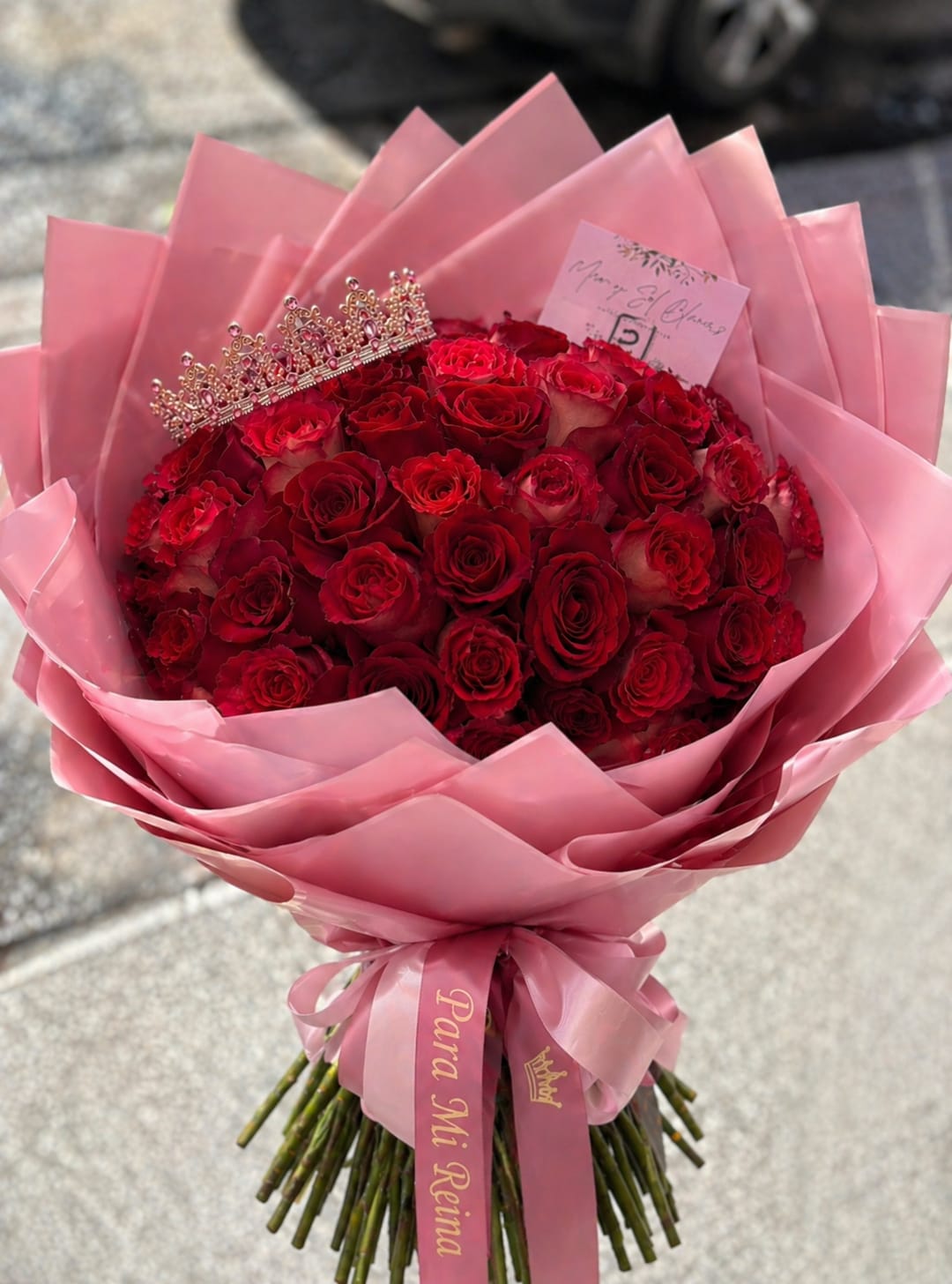 Mi Reina Rose Bouquet - Bold, romantic, and made to impress, this stunning bouquet features a lush dome of premium red roses, symbolizing deep love and admiration. Wrapped in elegant blush-toned layers and finished with a satin ribbon that reads “Para Mi Reina,” it’s crowned with a delicate tiara for that extra regal touch.