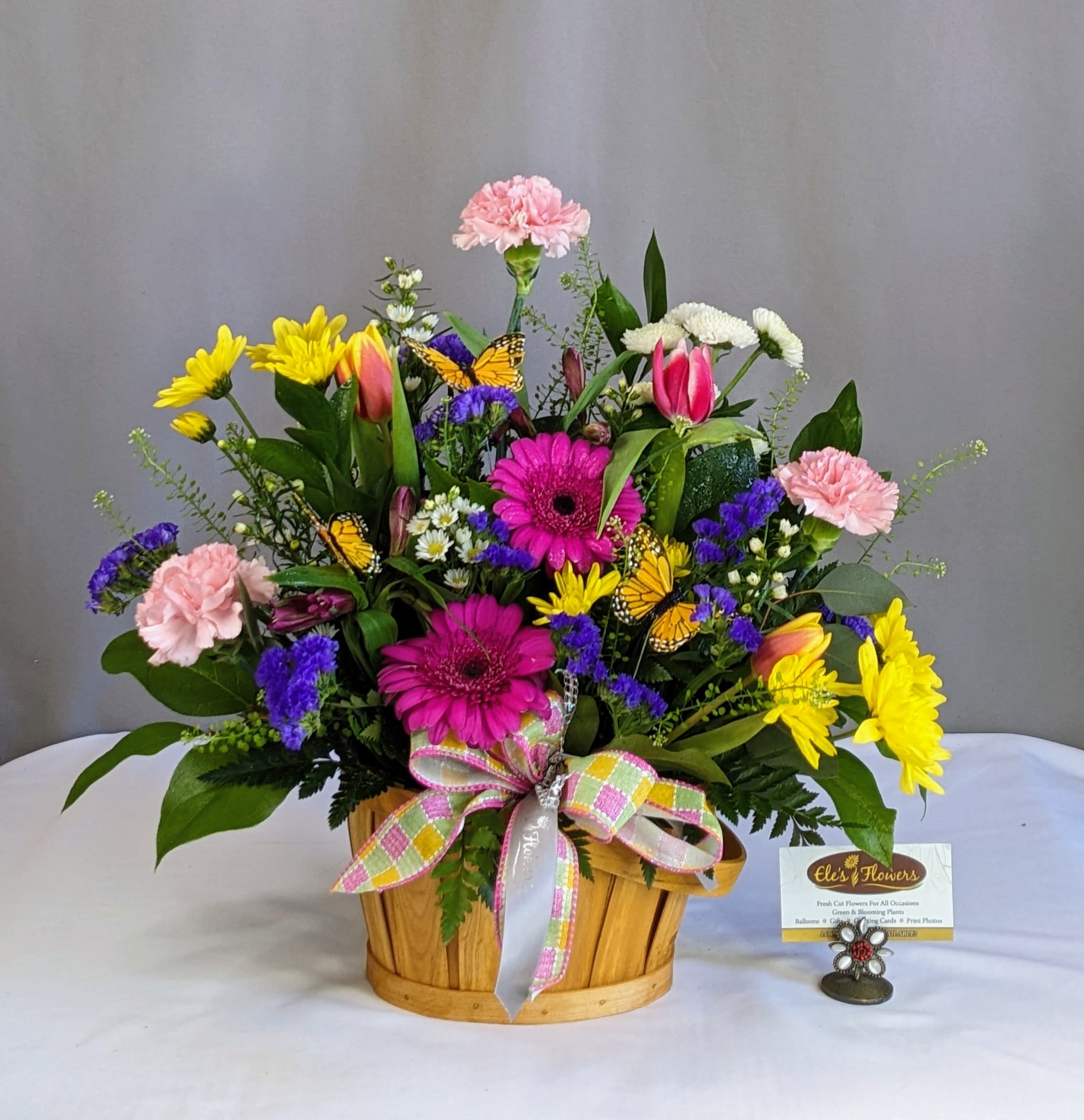 Jubilee Basket - Celebrate the joy of the season with our Jubilee Basket—a cheerful explosion of vibrant blooms that radiates warmth and happiness. This lively arrangement features a stunning mix of sunny yellow, pink and orange blooms, and pops of blue and purple flowers, all nestled in a charming wooden basket. Decorative butterflies bring a whimsical, garden-fresh feel, while a plaid pastel bow adds the perfect finishing touch. Perfect for any occasion, this arrangement is a burst of seasonal delight.   Blooms may vary from photo.    Standard Approximate Dimensions: 18"H x 17"W  Deluxe Approximate Dimensions: 20"H x 20"W