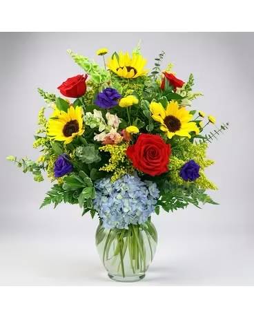 French Delight - Bright yellow sunflowers steal the show in this joyful flower arrangement, paired with red and purple roses for a rich, celebratory contrast-perfect as a birthday bouquet or a congratulations gift. The mix of pale blue hydrangea and delicate alstroemeria adds soft texture and a cool counterpoint to the warm tones, creating an upbeat, garden-style display that makes the person you're thinking of feel genuinely seen and celebrated.  Arranged in a clear glass vase, the design is full and layered with abundant greenery that frames each bloom and highlights the vibrant color palette of yellow, red, blue, and purple. The rounded vase gives a classic florist presentation and keeps the focus on the flowers' natural movement and texture; the varied sizes-from the sunflowers' bold faces to the hydrangeas' cloud-like clusters and the roses' velvety petals-deliver plenty of visual interest and presence for any table or desk.  Ideal for birthdays, congratulations, or simply sending a cheerful hello, this arrangement reads as both festive and thoughtful for many occasions. Standard care-fresh water in the vase and trimming stems every few days-will help the bouquet stay bright; order in time for the special day to ensure the recipient receives that immediate lift only fresh flowers can give.