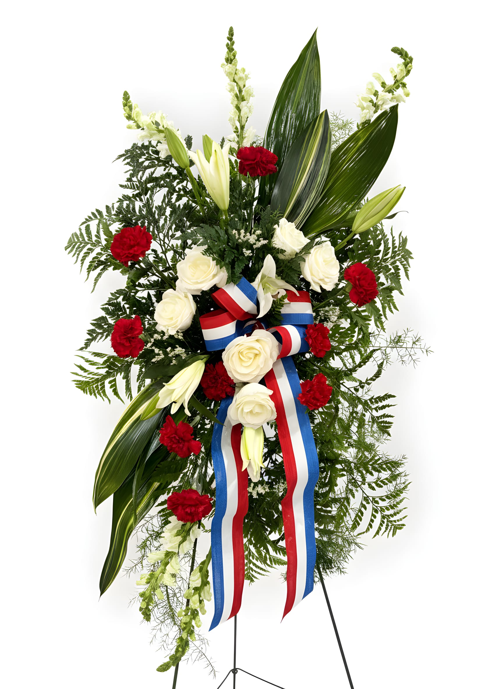 Patriot Stand spray - A tall, formal standing spray that reads with solemn dignity-perfect as a sympathy or funeral tribute and fitting for Memorial Day or Fourth of July observances. This Patriot Stand spray pairs creamy white roses and pale lily buds with bright red carnations and spray roses to create a respectful, vertical silhouette; the red, white and blue ribbon bow and streamers at the center give a clear patriotic accent without overpowering the arrangement.  Layered glossy foliage, fern-like textures and airy filler add depth and a ceremonial structure, so the arrangement carries well in a chapel, graveside setting, or memorial service. The mix of carnations, roses, lilies and spray roses produces a clear contrast against deep green leaves, offering a composed, reassuring presence that helps the person you're honoring feel remembered and respected.  Sized and mounted on a standing easel for easy display, this spray arrives ready to place at the service. Minimal care is needed other than keeping the base supplied with water until the time of the ceremony; order with confidence for a meaningful tribute that conveys honor, sympathy, and patriotic sentiment.